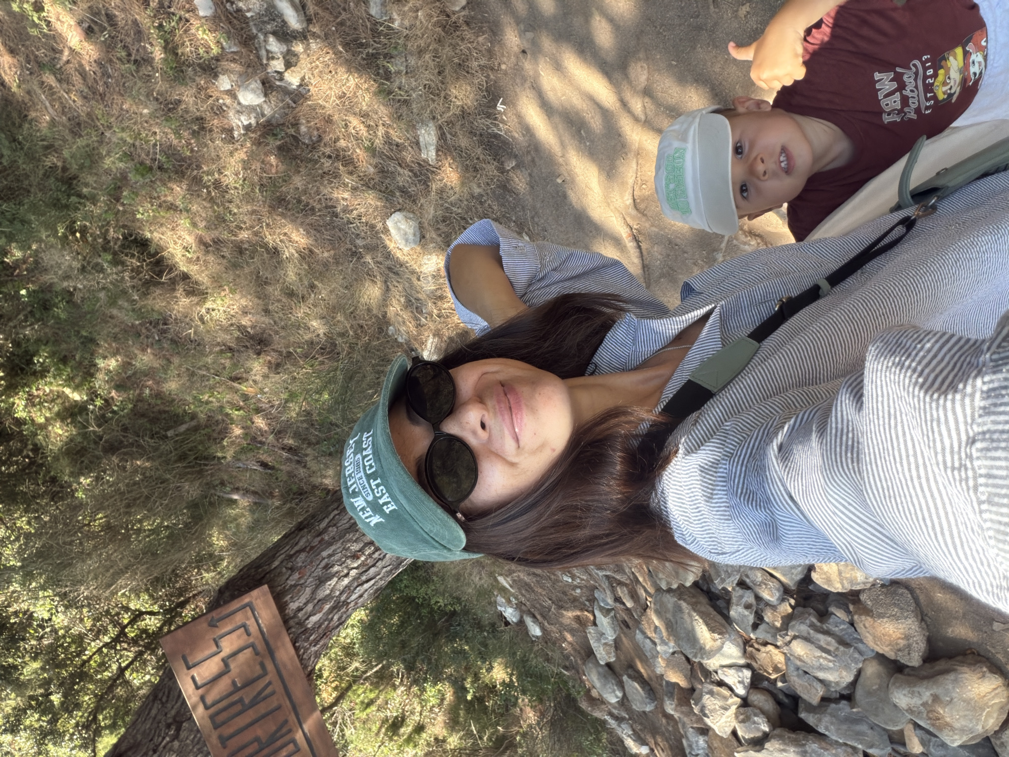A medium shot selfie shows a woman and a young boy outdoors in a sunny, wooded area. The woman, in the foreground, wears a green baseball cap that reads "NEW JERSEY EAST COAST," black sunglasses, and a blue and white striped shirt. She smiles at the camera, her right arm resting on her shoulder. To her left, a wooden sign nailed to a tree trunk has "ORTRESS" carved into it, with an arrow pointing upwards. A young boy, with light brown hair, is visible to the woman's right and slightly behind her. He wears a white baseball cap with green lettering and a maroon t-shirt with "PAW PATROL" graphics. The boy gives a thumbs-up to the camera and makes a slight grimace. The environment consists of a dirt path, scattered rocks, and dry grass, with trees and foliage in the background. The lighting suggests it is daytime. The scene is likely at the Mogren Fortress in Budva, Montenegro, given the "ORTRESS" sign.