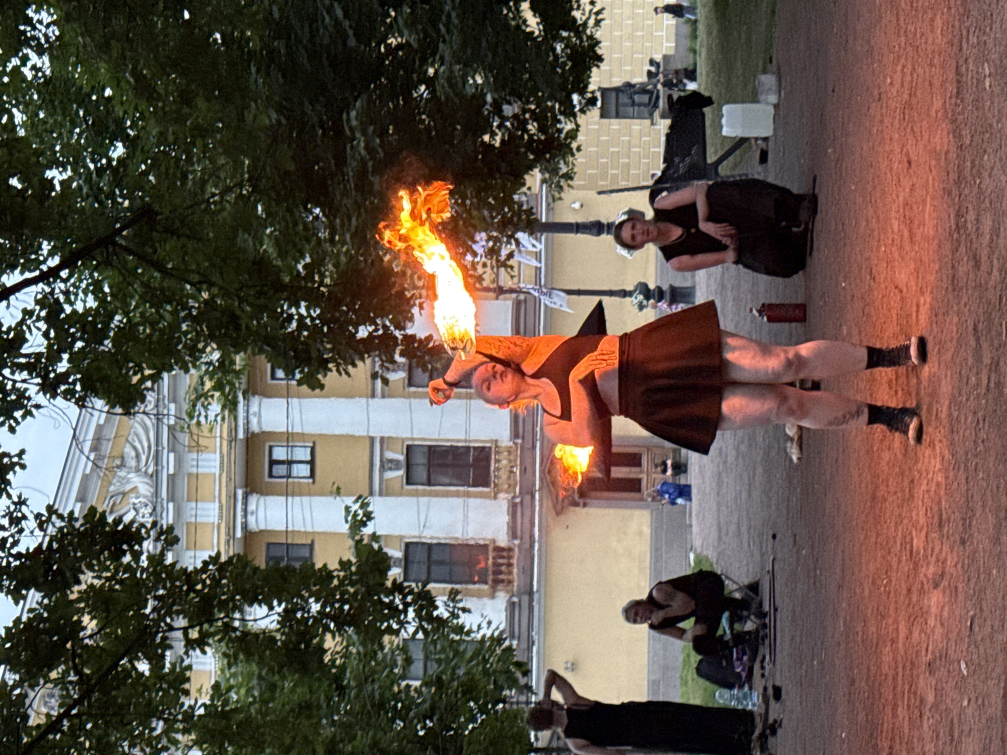 The image depicts a fire performance occurring outdoors in front of a building in Saint Petersburg, Russia. The central figure, a woman, is performing with fire, holding a flaming object in her right hand which emits a large plume of fire and smoke. She is wearing a dark crop top and a short dark skirt. She has tattoos on her arms and legs and is wearing dark shoes and socks.
In the background, a grand, pale yellow building with white columns and classical architectural details is visible. Trees with dense green foliage partially obscure the building. Several other individuals are present in the scene. To the left, a person in a black dress stands with their back to the camera. In the middle ground, two people are seated on the ground. One, a person with blonde hair, is sitting on a small stool and looking down. Another person, dressed in dark clothing, is kneeling and looking towards the viewer. Various objects, including bags, bottles, and what appears to be a fire extinguisher, are scattered on the ground. The ground is a reddish-brown gravel or dirt path, illuminated by the orange glow of the fire. The overall atmosphere suggests an outdoor performance or gathering.