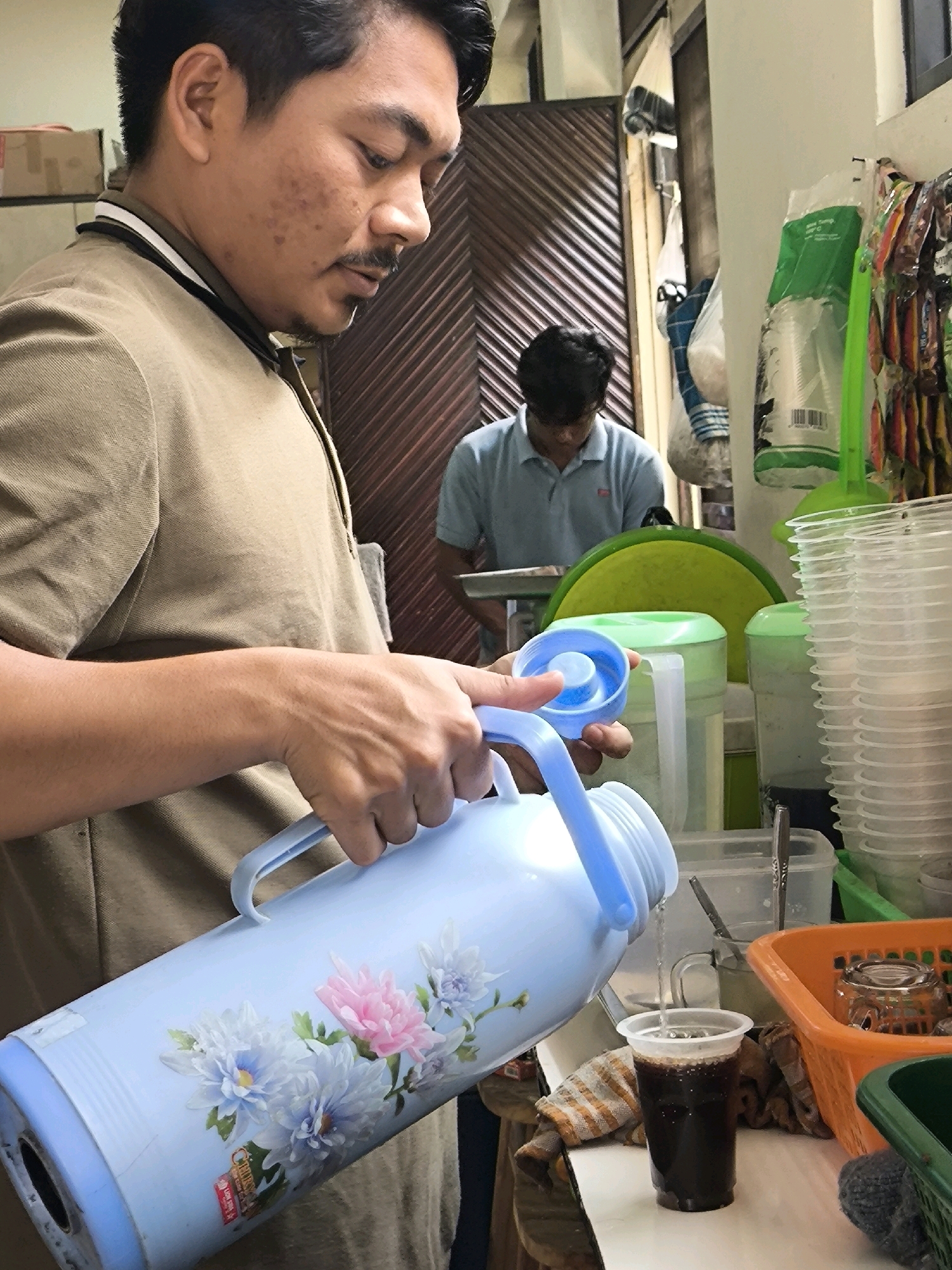 The image captures a close-up, low-angle view of a man in West Jakarta, Indonesia, pouring a dark liquid from a large, light blue thermos with floral decals into a clear plastic cup. The man, wearing a tan collared shirt, has dark hair and a mustache and is looking down at the cup, focused on his task. His right hand holds the thermos handle, while his left hand is near the spout. In the background, another person in a light blue polo shirt is visible, partially obscured by the foreground action. The setting appears to be an indoor space, possibly a shop or a home kitchen, with various items like stacked plastic cups, containers, and shelving visible. The overall lighting is moderate, and the scene suggests an everyday moment of serving a beverage.