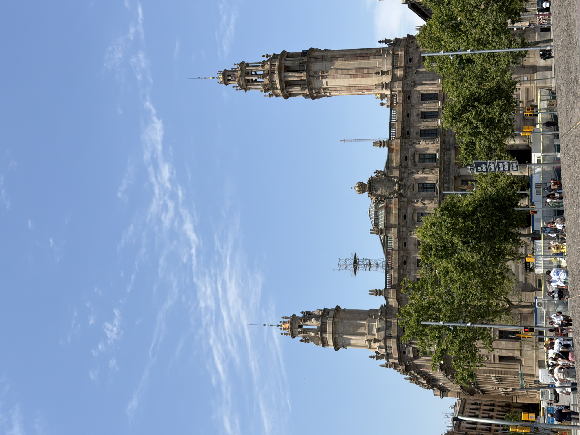 The image shows a large, ornate building with two prominent towers, one on the left and one on the right. The building is made of stone with intricate architectural details, including statues and decorative moldings. The towers are tall and cylindrical, with multiple levels and spires. The building appears to be situated in a public square, with trees lining the front and people walking around. The sky is blue with some scattered clouds.
The building in the image is the Palau de la Virreina, located on La Rambla in Barcelona, Spain. It is a Baroque palace built in the 18th century and is known for its impressive architecture and its role in the city's cultural life. The Palau de la Virreina is a popular tourist attraction and a significant landmark in Barcelona. The image captures a sunny day, with people enjoying the outdoor space in front of the palace.