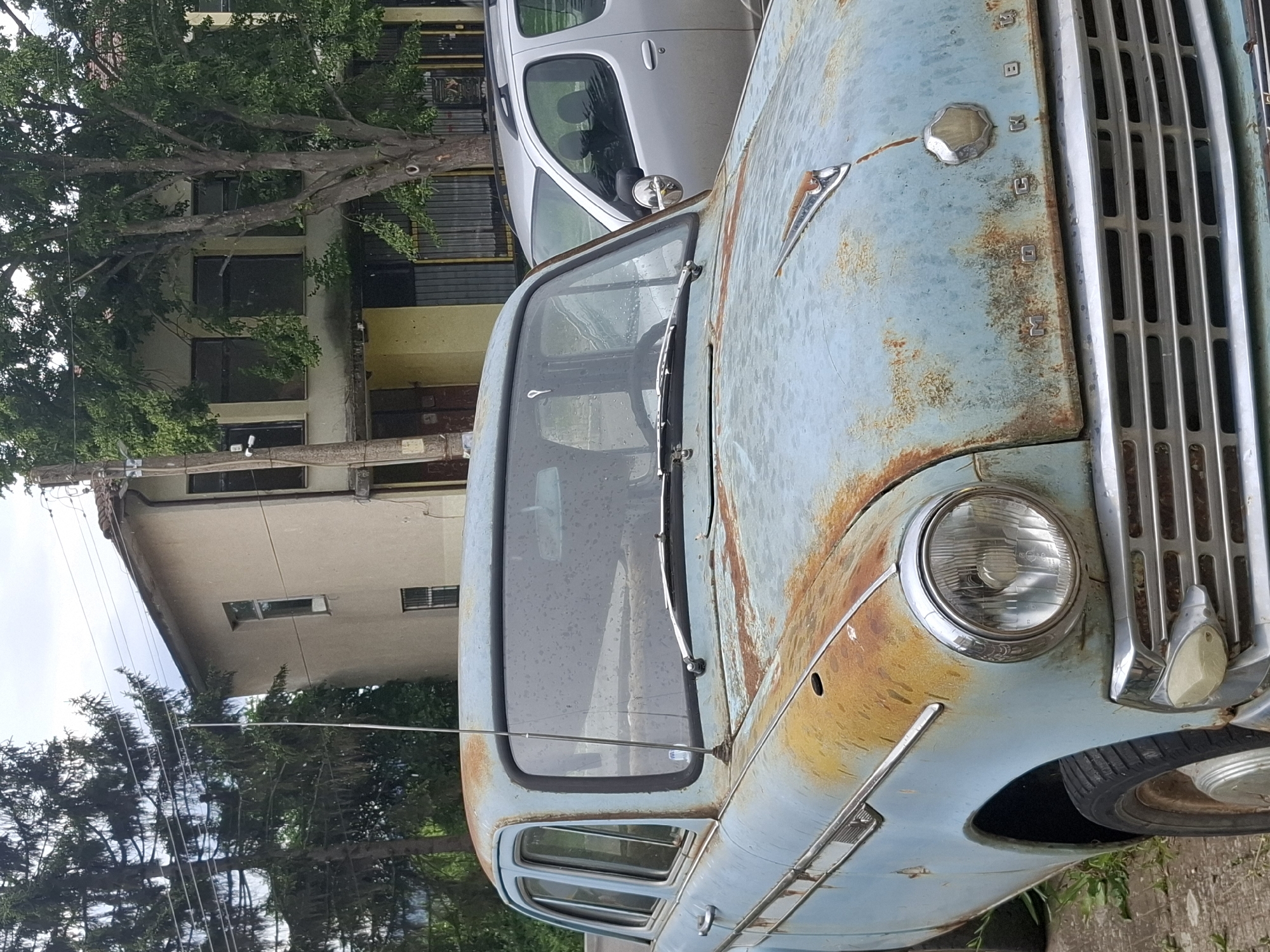 The image shows the rusted front of an old, blue car with a prominent chrome grill. "BUCKINGHAM" is vertically spelled out on the vehicle next to the grill. The windshield is cracked. The car appears to be parked on a street in the city of Baylovo, Bulgaria, as indicated by the architecture of the adjacent buildings and the presence of trees in the background. Another modern, silver car is parked nearby. There are no people visible in the immediate frame. Power lines run through the image as well.