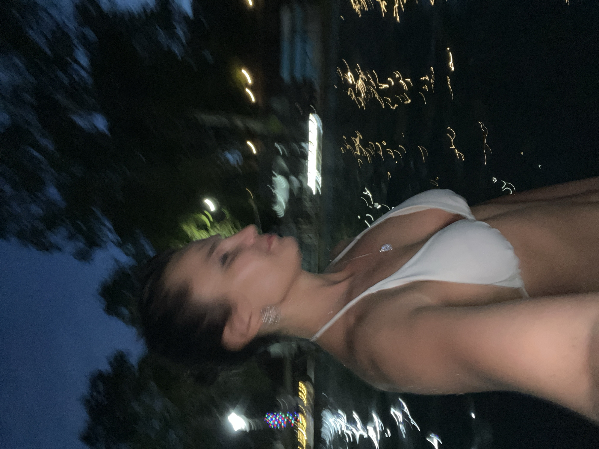 The image depicts a woman in a white bikini at night. She is positioned on the left side of the frame, facing right in profile, with her head turned slightly away from the camera. Her dark hair is pulled back. She is wearing a necklace with a pendant and earrings. The background shows a body of water with reflections of lights, suggesting an outdoor setting at night. Trees and foliage are visible in the darkness behind the water. The overall lighting is dim, consistent with nighttime. The scene is not specifically identifiable as being in Paratunka, Russia, as there are no unique landmarks or contextual clues to confirm this location.
