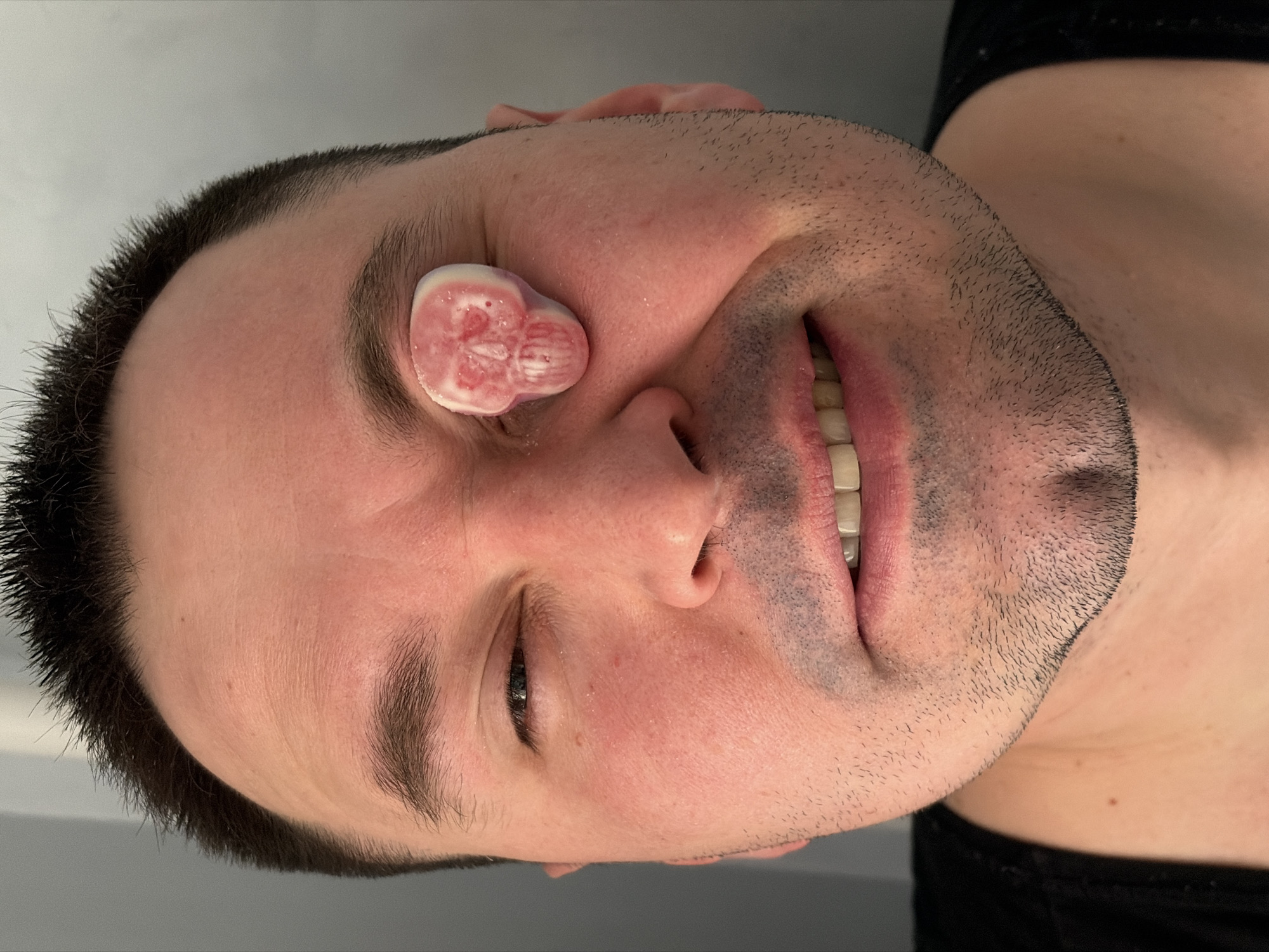A man's face is visible, turned sideways to the camera. He has short, dark hair and light skin. One eye is closed and the other is partially open, revealing a dark iris. The man is smiling slightly, exposing his teeth. A white candy in the shape of a skull with pink details rests on his face, near his eye. He is wearing a dark shirt. The background is a plain light gray. No location in Vozhoiskii, Russia is indicated in the image.