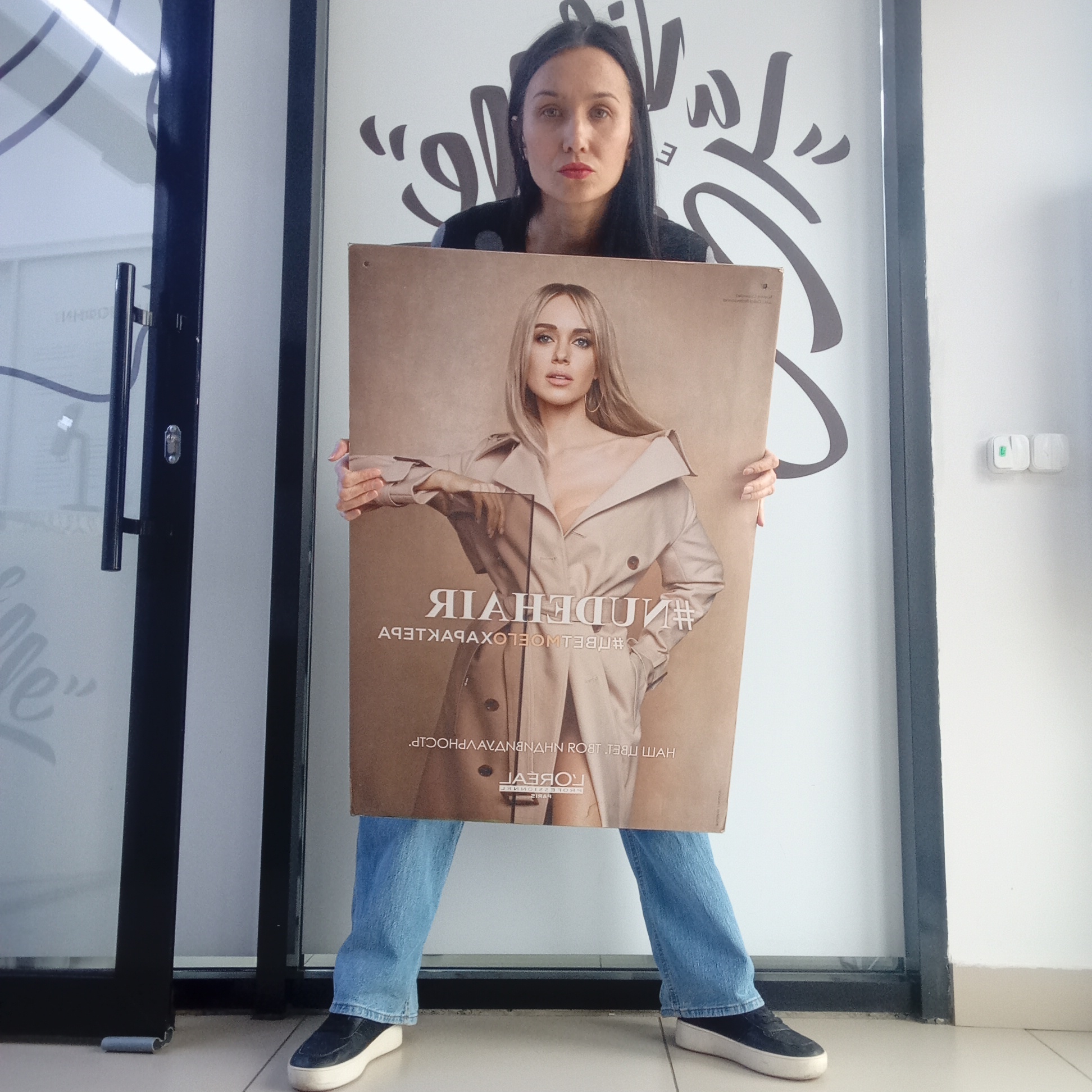 A woman with dark hair and red lipstick stands indoors, holding a large, inverted advertisement poster featuring another woman. She is dressed in a dark top, blue jeans, and black platform sneakers with white soles. Her expression is neutral as she holds the poster, which largely obscures her body from the chest down.
The poster displays a blonde-haired woman with blue eyes, wearing a beige trench coat. The text on the poster is reversed, indicating it might be designed for reflection or viewing through a transparent surface. When viewed correctly (mirrored), prominent elements include the hashtag "#NUDEHAIR" and the "L'ORÉAL" logo. Other reversed Cyrillic text is also visible on the poster.
The setting appears to be an indoor commercial space, possibly a salon or shop, in Izhevsk, Russia. Behind the woman is a glass door with a black frame and a silver handle, revealing a faint reflection of text. To the right of the door is a white wall with dark grey, stylized graphic art or text. Further to the right, on a plain white wall, is a white electrical outlet or light switch. The floor is light-colored tiles. The woman seems to be posing for a photograph while displaying the product advertisement.