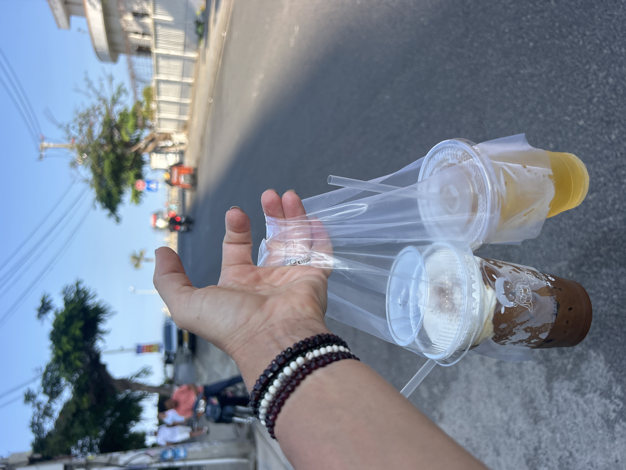 A first-person perspective photograph captures a person's hand holding two takeaway drinks in a clear plastic carrier bag, set against a bustling outdoor street scene. The hand, adorned with multiple beaded bracelets in dark red/brown and white, along with a ring on the middle finger, extends into the foreground. One drink is a light yellow or orange liquid, possibly a tea or juice, while the other contains a dark brown liquid, likely coffee or chocolate, topped with a generous layer of white cream or foam. Both plastic cups have clear domed lids and are accompanied by plastic straws.
The background is a sunlit street, consistent with Bắc Nha Trang, Vietnam. The asphalt road stretches into the distance, flanked by light-colored buildings with various architectural details, including fences and grates. Lush green trees are scattered along the roadside, partially obscuring some structures. The sky above is clear and bright blue, suggesting a sunny day, likely late morning or afternoon given the strong lighting. In the mid-ground, a red motorbike or scooter is visible on the street, and other blurry vehicles can be discerned further back, including what appears to be a white vehicle. A utility pole with overhead wires is also present. A faint, stylized white logo or graphic is visible on the side of the dark brown drink cup, but any accompanying text is indistinct and unreadable. The scene conveys a casual moment of getting refreshments while out and about.