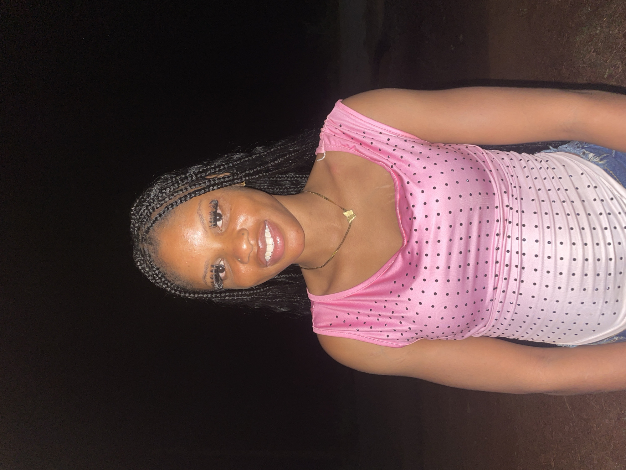 A dark-skinned woman with braids is smiling at the camera. She is wearing a pink and white polka-dotted dress and a gold necklace. The background is dark, suggesting it is nighttime. The location is the city of Ogbe, Nigeria.