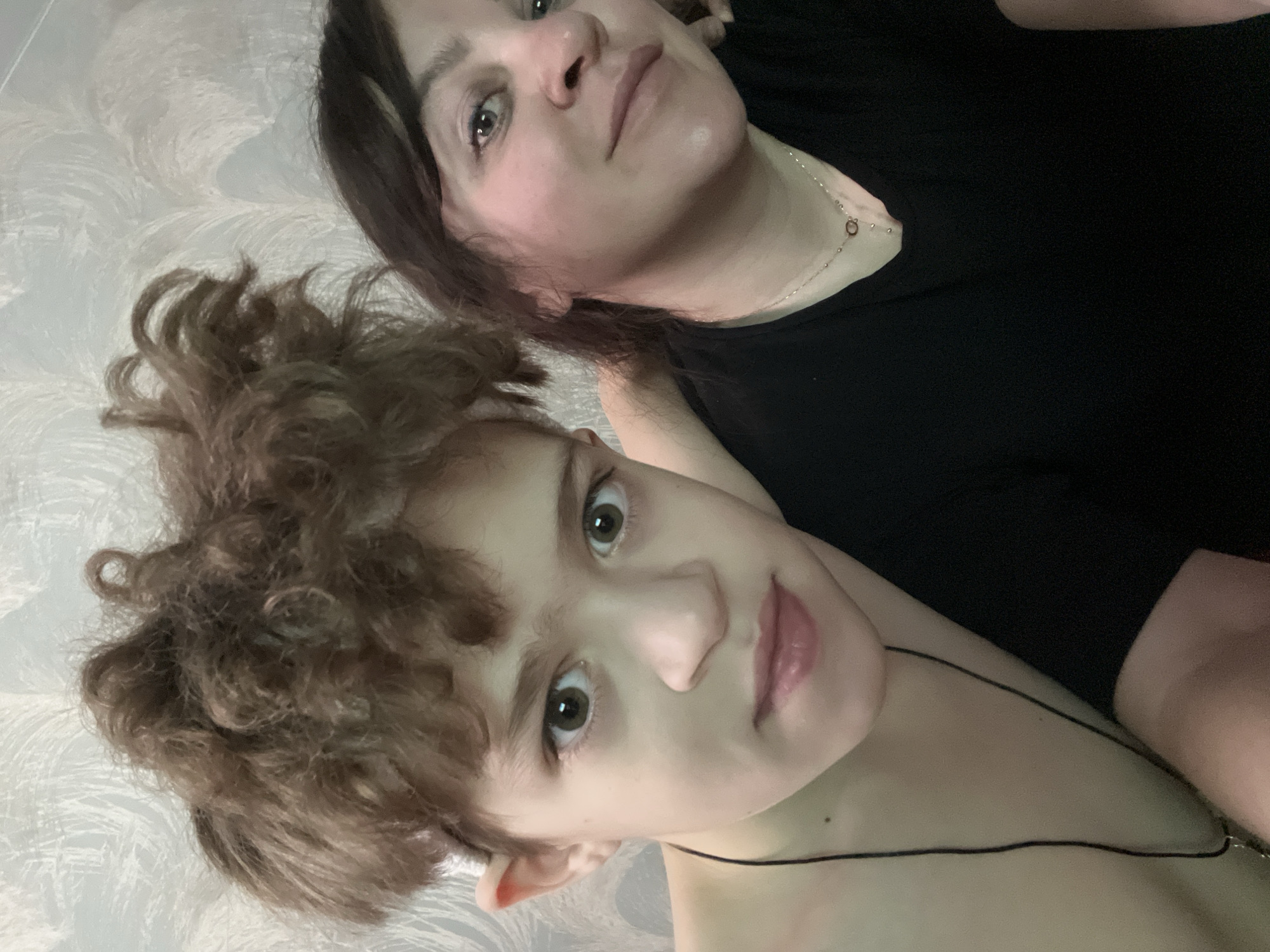 An indoor portrait displays two individuals: a male youth positioned prominently in the foreground, and an adult female partially visible behind him. The male youth, with light brown, curly hair and wide brown eyes, looks directly at the camera with a neutral expression. He appears shirtless, wearing a thin black cord necklace. The adult female, positioned to the right and slightly behind, has dark brown hair and offers a subtle smile towards the camera. She wears a black v-neck top and a delicate silver-colored chain necklace featuring a small, light-colored circular pendant. The background consists of a wall covered in light-toned wallpaper displaying a subtle, abstract, feather-like pattern. No visual information within the image indicates the specific city of Noviy, Russia.