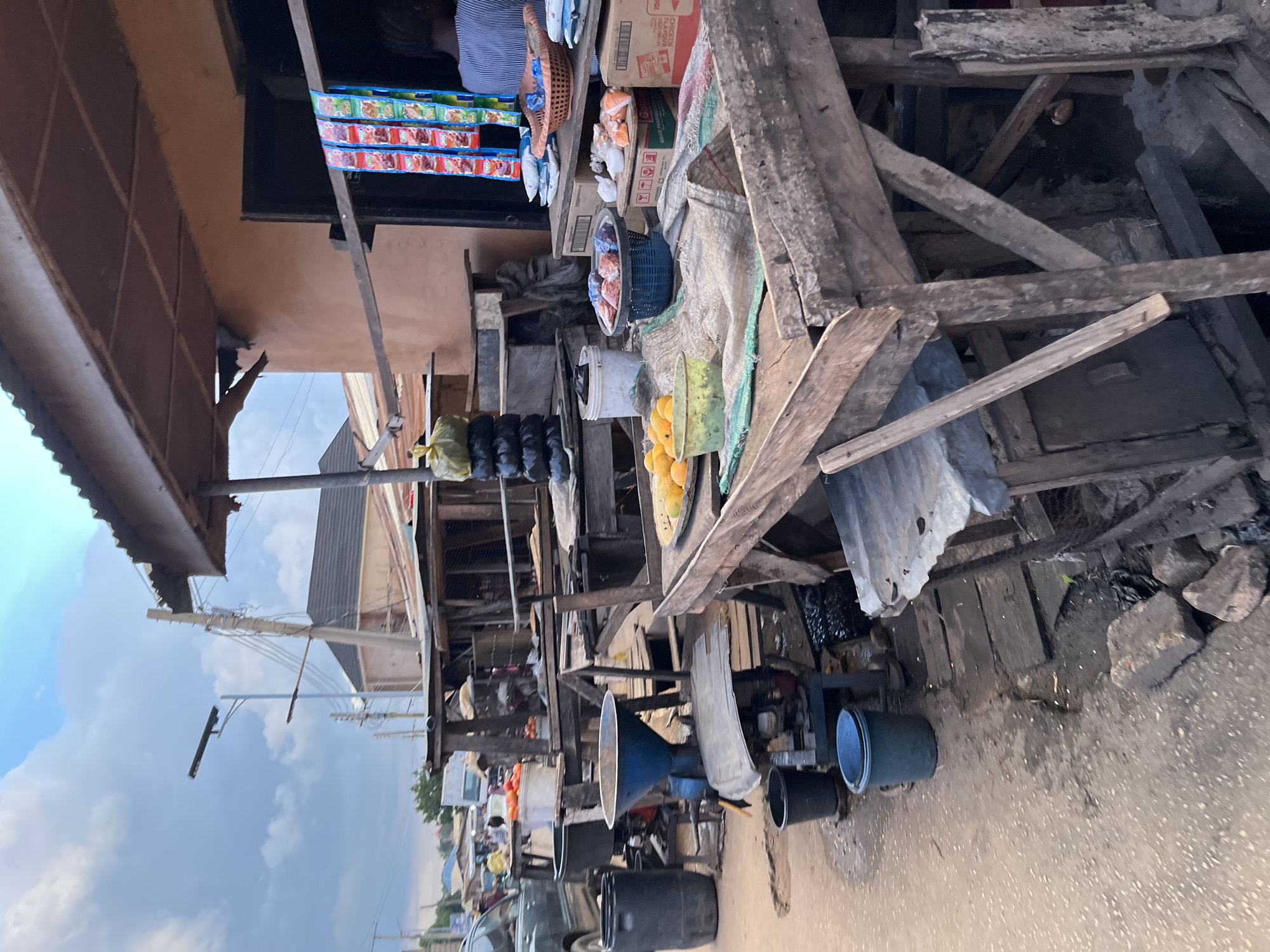 This image captures a vibrant, cluttered street market or roadside stall during the daytime, viewed from a slightly elevated perspective. The scene is characterized by makeshift structures constructed from rough wooden planks and beams, supporting corrugated metal sheets that likely serve as roofing or shelving. The ground below is unpaved earth, possibly dusty or muddy in parts, with scattered rocks and debris.
In the upper left quadrant, partially obscured by the stall's roof, a person wearing a striped blue and white shirt is visible, likely a vendor. Below them, rows of colorful snack packets are prominently displayed, hanging from a beam.
The stall is laden with various goods. To the right of the vendor, cardboard boxes, one partially labeled "CHICKEN FOOD," are stacked. Further along the stall, a blue woven basket holds an assortment of items, and a green plastic basin is filled with numerous yellow and orange fruits, possibly mangoes or oranges. Other plastic buckets, some black and some white, are scattered around the display area and on the ground. Several black items, possibly plastic bags or smaller goods, hang near the fruit.
Prominently featured in the lower right and center is a large, light blue, conical funnel, appearing to be part of a larger piece of machinery, possibly an agricultural mill or processing unit. Nearby, another piece of equipment with engine-like components is visible.
In the background, behind the stall structures, utility poles and electrical wires are discernible against a cloudy sky, suggesting an open, outdoor urban or semi-urban environment. The overall impression is one of bustling commerce in an improvised setting.