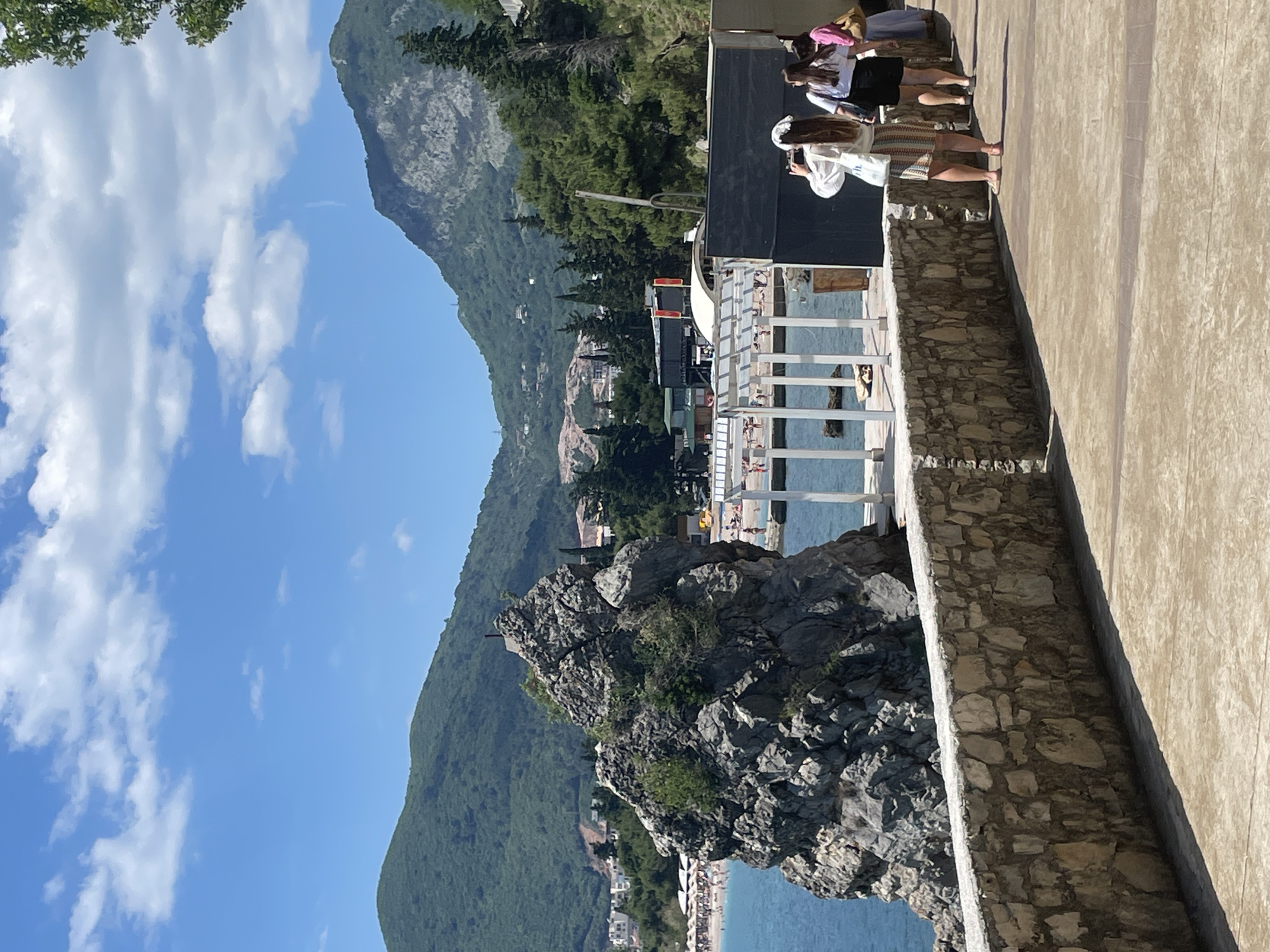 The image captures a sunny, outdoor scene from Budva, Montenegro. A group of people stands on a stone-walled promenade overlooking a rocky coastline with a beach below. Some people are taking pictures. The background features mountains. The sky is blue with scattered clouds. The structures and vegetation imply a coastal resort environment.