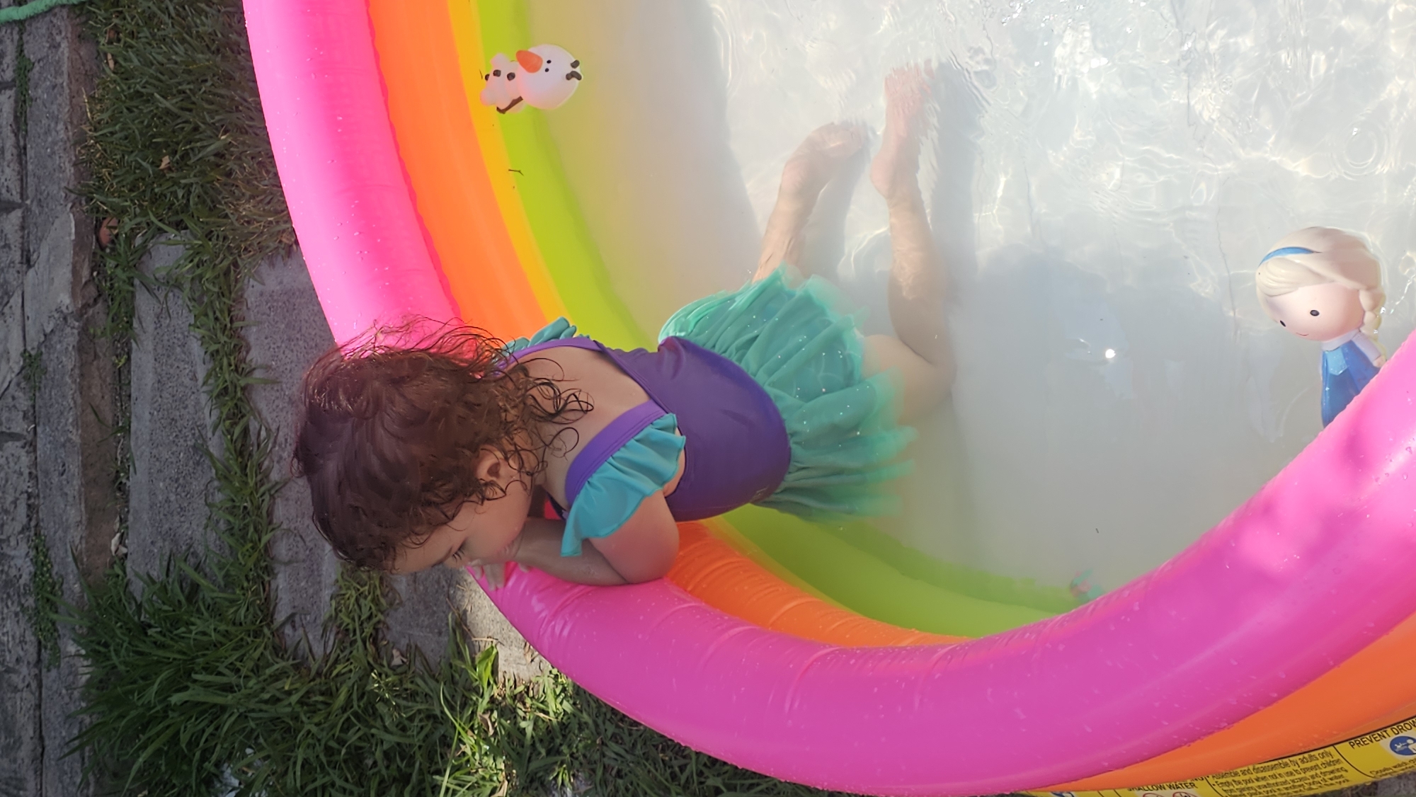 The image shows a young child, likely a girl, in a brightly colored inflatable pool with multiple colors, including pink, orange, green, and yellow. She is wearing a purple and turquoise swimsuit with a ruffled skirt. The child is partially submerged in the clear water, with her feet visible and facing upward. The pool also contains two floating toys: one is a small white figurine of Olaf from the "Frozen" movie franchise, and the other is a doll of Elsa from "Frozen". The pool is surrounded by grass on one side and pavement or concrete on the other. The lighting suggests it is daytime. The child appears to be relaxing or playing in the water. This scene is located in San Bernardino Tlaxcalancingo, Mexico.