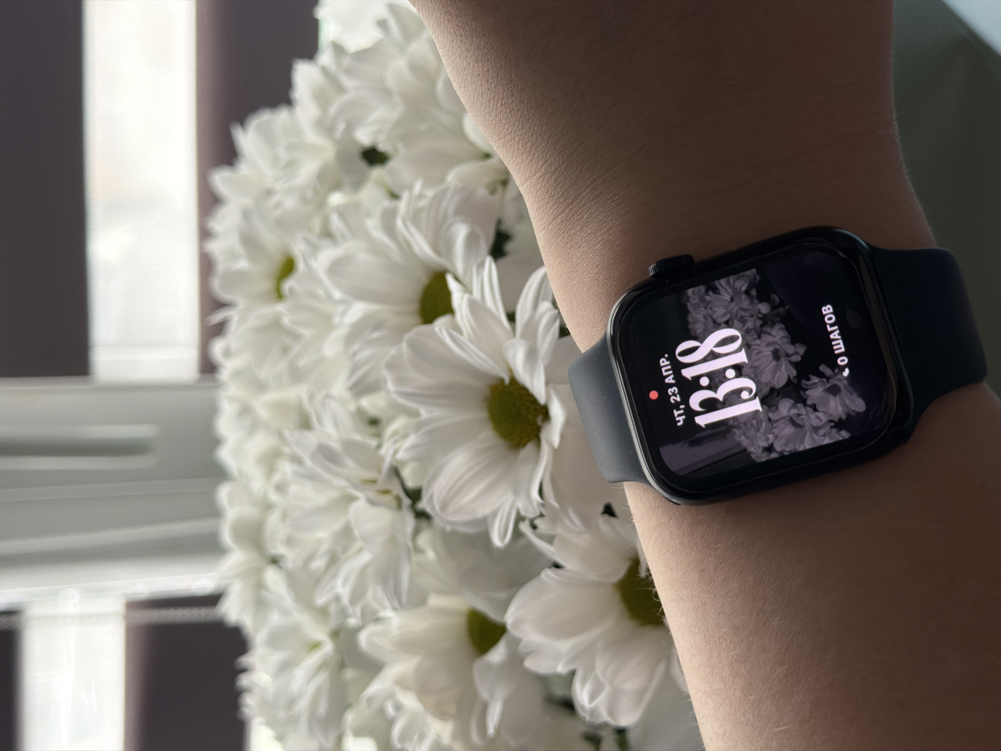 An eye-level close-up shot features a person's fair-skinned arm wearing a black Apple Watch, positioned in the lower right of the frame. The watch face is brightly lit, displaying Russian text. It shows "ЧТ, 23 АПР." (Thursday, April 23) and the time "13:18". A floral pattern, similar to the real flowers in the background, is visible on the watch face, along with "0 ШАГОВ" (0 steps). The watch has a dark band.
Behind the arm and watch, a large bouquet of white flowers, likely chrysanthemums or daisies, is in soft focus, filling the left and central parts of the image. Their numerous petals create a soft, textured backdrop. The far background is blurred, revealing vertical elements that appear to be window blinds or curtains, indicating an indoor setting with natural light source. The lighting is bright and even, consistent with early afternoon. Visible details on the arm include fine hair and natural skin texture. The Russian text on the watch aligns with a location such as Chaikovsky, Russia.