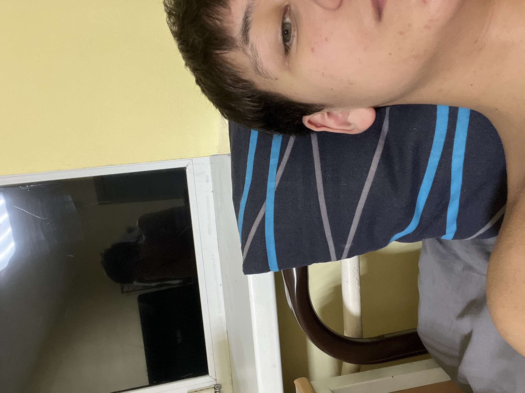 The scene depicts an interior view of a room, likely a private residence, located in Kazan, Russia. A person, identified by a partial view of their head and upper torso, is positioned on the right side of the frame, lying down. The individual has short, dark hair and light-colored eyes. Their face exhibits minor skin blemishes. The person is resting their head on a dark blue pillow adorned with light blue and grey horizontal and diagonal stripes. A grey sheet or blanket is visible beneath the pillow.
Behind the person and pillow, a curved, dark brown wooden chair back is partially visible. In the upper left quadrant of the image, a white-framed window or partition is present. The dark, reflective surface of a screen, possibly a television or monitor, occupies the upper central area, reflecting the room. This screen displays noticeable scratches. Within the reflection on the screen, the individual's shirtless upper body and head are clearly visible, confirming their position relative to the screen. A black laptop, with faint lettering resembling "ALURA" on its lid, is also reflected on the screen, positioned beneath the person's reflection. The walls of the room are painted a muted yellow color. The lighting within the room appears to be artificial, casting reflections on the screen's surface.
