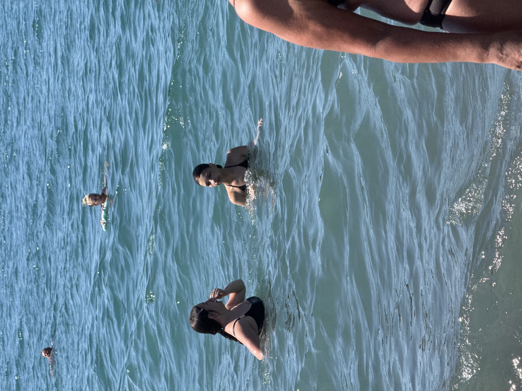 A group of people are swimming in the blue waters of the Black Sea, off the coast of Sochi, Russia. The water is choppy with small waves. In the foreground on the right, a person's arm and torso are visible, wearing a black bikini bottom. In the middle ground, two young women are swimming. The woman on the left is facing towards the left and appears to be touching her face. The woman on the right is facing forward, looking towards the left. In the background, a woman with blonde hair is swimming with a green float, and a man is also visible swimming further in the distance. The sky is clear and the sun is shining.