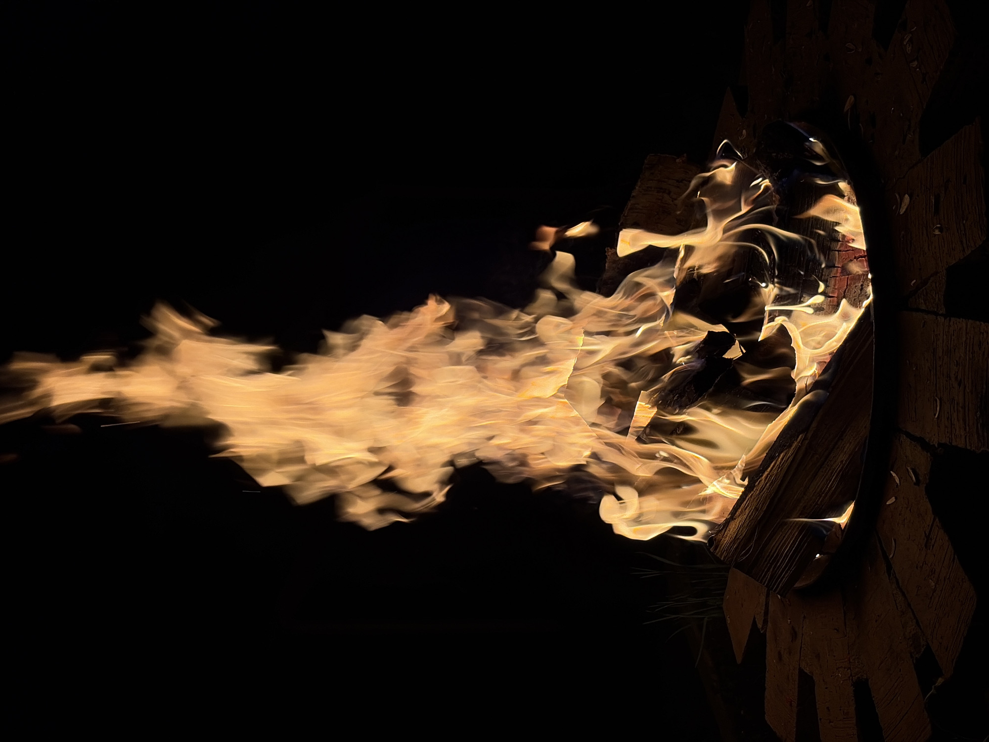A long exposure image shows a fire in a hearth constructed of stacked wooden blocks in Malyi Bukor, Russia. The flames extend from the hearth to the left and appear blurred, suggesting motion over a short amount of time. The wooden blocks are a dark brown color and are arranged to create a circular opening through which the fire can be seen. The background is dark.