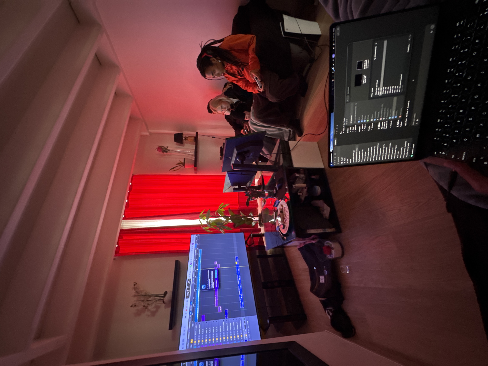 A low-angle, indoor shot shows two individuals sitting on a dark couch or futon. One individual has long dreadlocks and wears an orange hooded jacket. The other person has light blonde hair and wears dark clothing. They appear to be looking at or interacting with a handheld electronic device. A black laptop computer, seemingly a MacBook Pro, is positioned in the foreground. Its screen displays programming or audio editing software. In the midground, there is a multi-screen computer setup with at least one monitor displaying an audio editing timeline. The room is lit with a reddish hue. Behind the computer monitors, red curtains partially cover a window. A small bookshelf with decorative items is also visible. The location is unlikely to be Paris, France, as there are no specific architectural or environmental features in the photo to confirm that the image was captured in Paris, France.