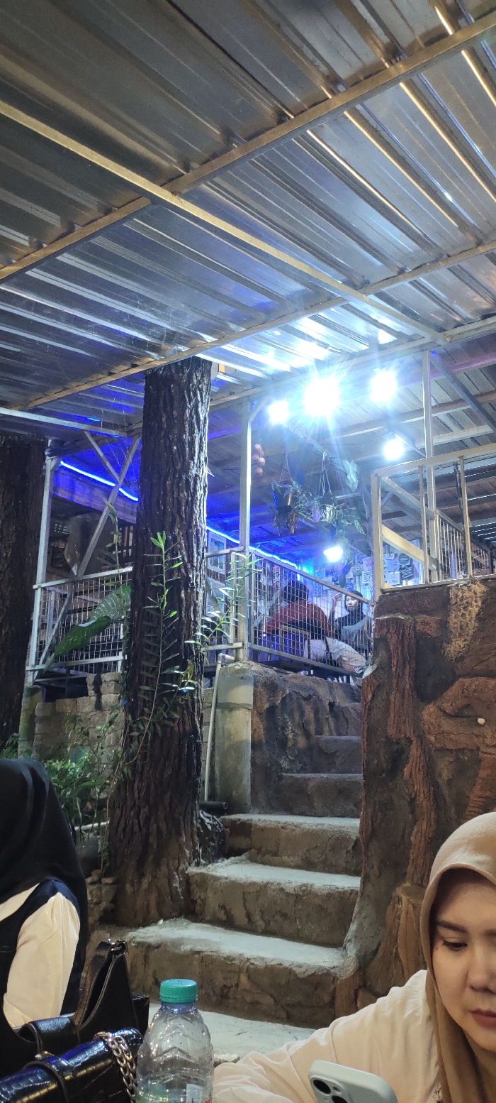 The image depicts a multi-level outdoor or semi-outdoor establishment, identified as being in Cembor, Indonesia, under artificial lighting, suggesting a nighttime or low-light setting.

The foreground features a surface, likely a table, on which a clear plastic water bottle with a blue-green cap is positioned. To the left, the shoulder and arm of an individual wearing a dark top and a white-and-blue garment are visible, alongside a black handbag with a silver chain strap. To the right, a woman with a light complexion wears a light brown hijab and a light-colored long-sleeved top. Her face is partially obscured by a light-colored smartphone she holds in her left hand, with her gaze directed slightly away from the camera.

In the midground, a series of stone or concrete steps ascends towards an elevated platform. A large, rough-barked tree trunk acts as a central structural column, with sparse green foliage growing on its bark. Another tree trunk is partially visible on the far left. A white PVC pipe is affixed vertically near the base of the central tree trunk.

The elevated platform in the background is enclosed by metal railings. Several individuals are seated behind these railings; one person in a red top is identifiable on the left side of the group, and another individual with dark hair is visible further to the right. The platform features decorative elements resembling rock formations.

Overhead, a corrugated metal roof is visible, supported by metal beams. Multiple bright white light fixtures are mounted to this structure, emitting strong glare and reflections. Blue LED strip lighting illuminates sections of the upper level. Hanging potted plants and what appear to be decorative gourds are suspended from the ceiling structure. Various papers or posters are affixed to a wall section on the upper right side of the elevated platform.