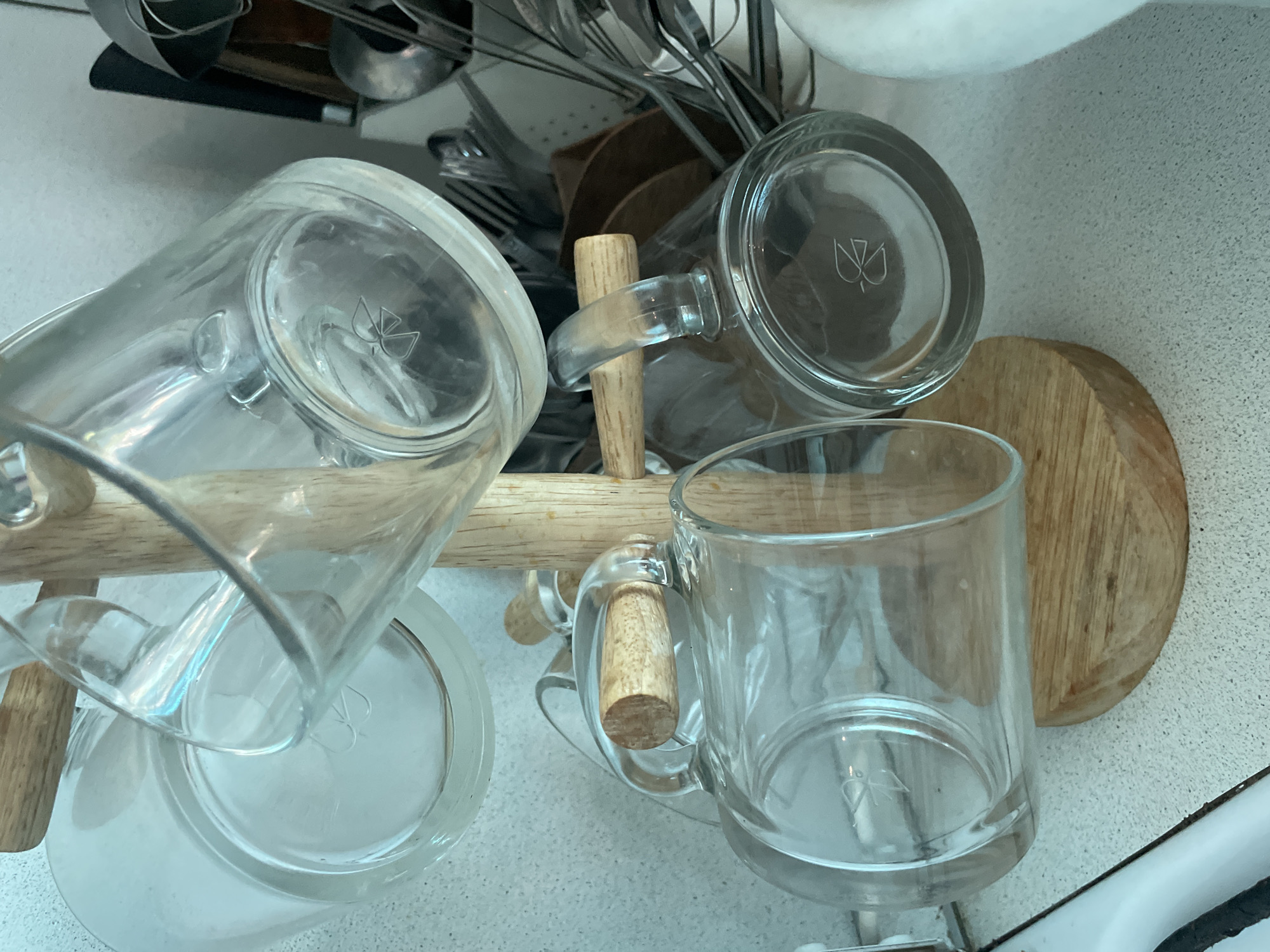 The image depicts three clear glass mugs with wooden handles inverted on a wooden mug tree. The mugs each have a logo on their base. Behind the mugs is a collection of kitchen utensils. The scene appears to be a kitchen counter, possibly in Olginka, Russia, but there is no definitive visual evidence in the image to confirm that location.