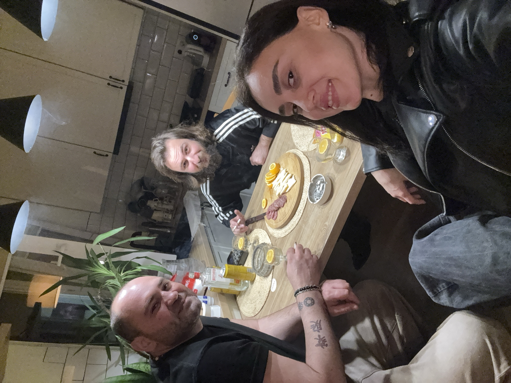 The image depicts three people sitting around a table in an interior space, likely a kitchen. One person, a woman with dark hair and wearing a black leather jacket, is taking the photo. The man to her left has long hair and a beard, wearing a black Adidas track jacket. The third person, a bald man wearing a black t-shirt with tattoos on his forearm, is positioned at the bottom of the image.
The table is set with food and drinks, including a wooden platter with cheese, orange slices, and salami, as well as several bottles and glasses.
The background includes kitchen cabinets, tiled walls, and pendant lights. A green plant is also visible.
The city of Tbilisi, Georgia, cannot be confirmed from this image.