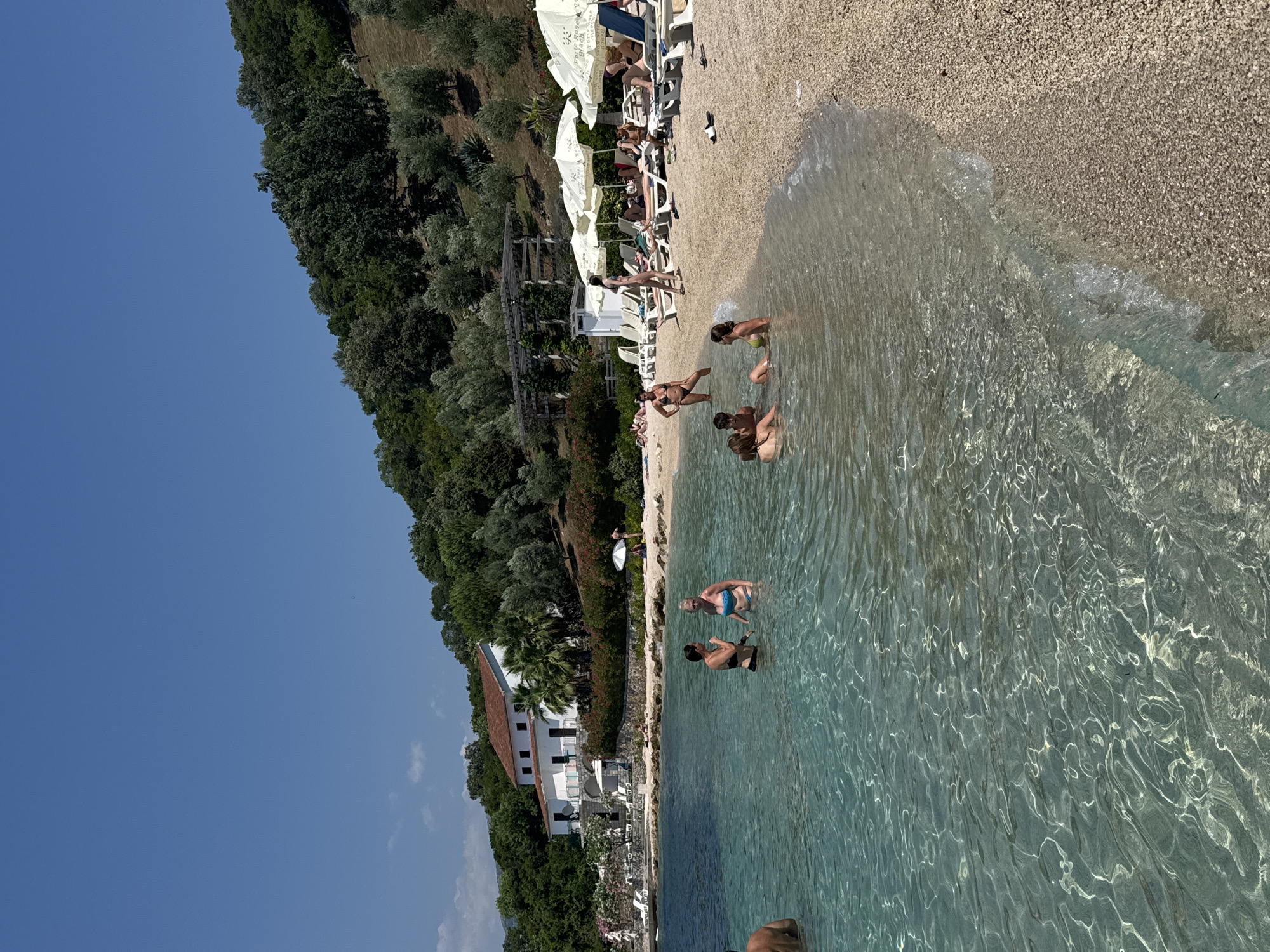 A high-angle, wide shot captures a sunny beach scene with a body of clear, turquoise water in the foreground. The water is dotted with several people swimming and wading. To the right, a pebble beach is lined with white lounge chairs and umbrellas, with more people sunbathing and relaxing. In the background, a white building with a red roof is nestled amongst lush green trees on a hillside. The sky is a clear, bright blue.
