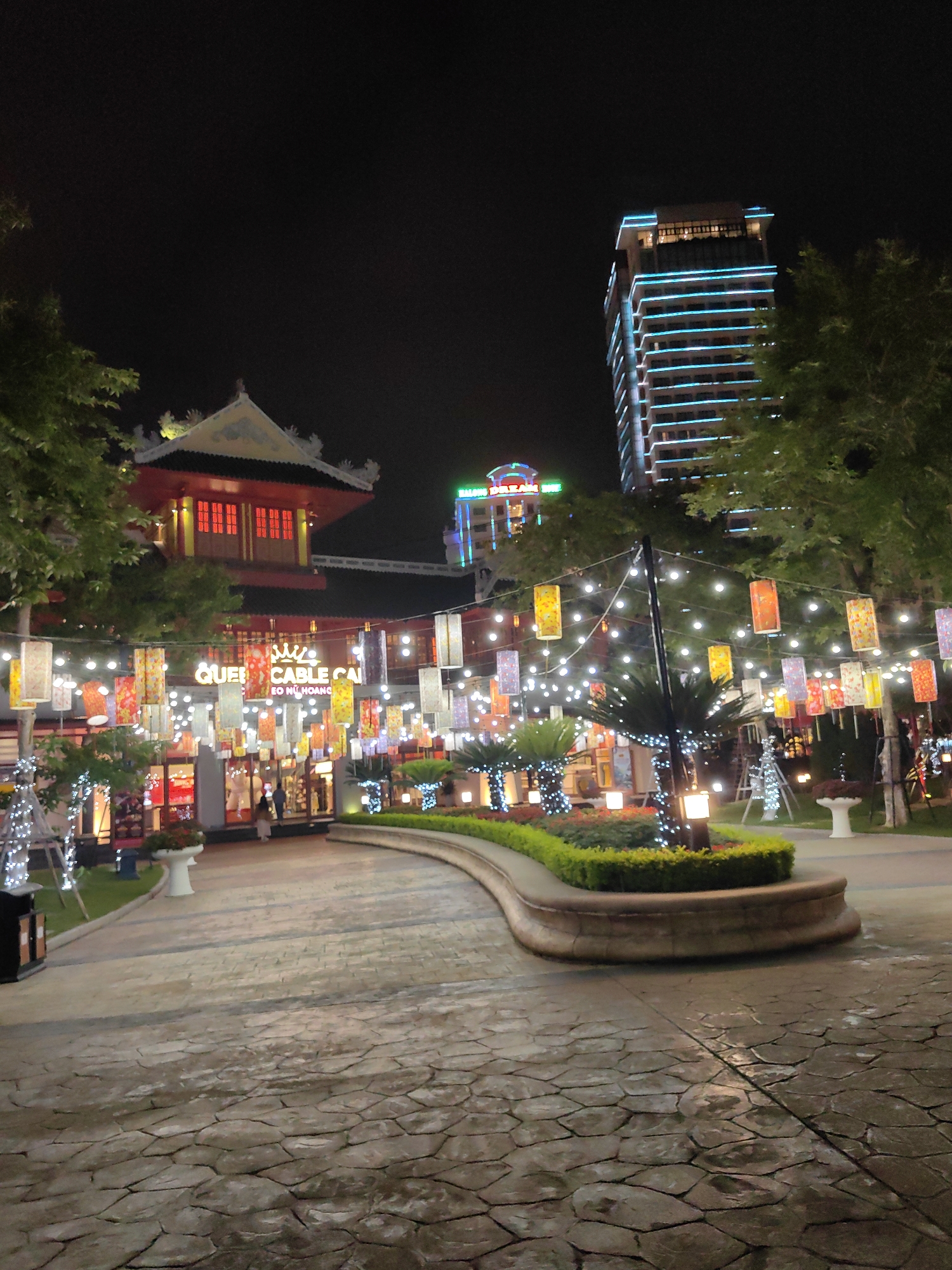 Captured at night, the image presents a scene in Bai Chay Ward, Vietnam. The foreground features a stone-paved pathway leading towards a lit building with a sign reading "QUEEN CABLE CAI." Overhead, strings of colorful lanterns illuminate the path. Vegetation is observed on either side of the walkway. In the background, multiple buildings rise, with one prominently displaying blue lights. The sky is dark, contrasting with the numerous artificial light sources in the scene.