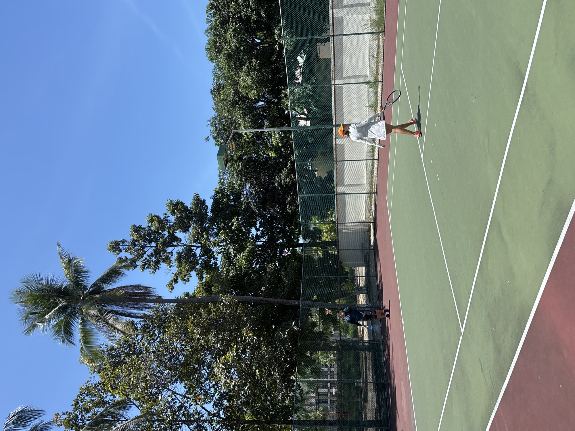 The image shows an outdoor tennis court with two people playing tennis. The court is green with red outer areas and white lines. In the foreground, a person wearing a white tennis outfit and an orange hat is walking away from the camera, holding a tennis racket. In the background, a person wearing a dark blue shirt and black shorts is standing on the court, also holding a tennis racket. The court is surrounded by a green fence and trees, including a prominent palm tree. The sky is clear and blue. The scene appears to be in a tropical or subtropical location. This appears to be a tennis court at a resort in Nha Trang, Vietnam, likely during the daytime.