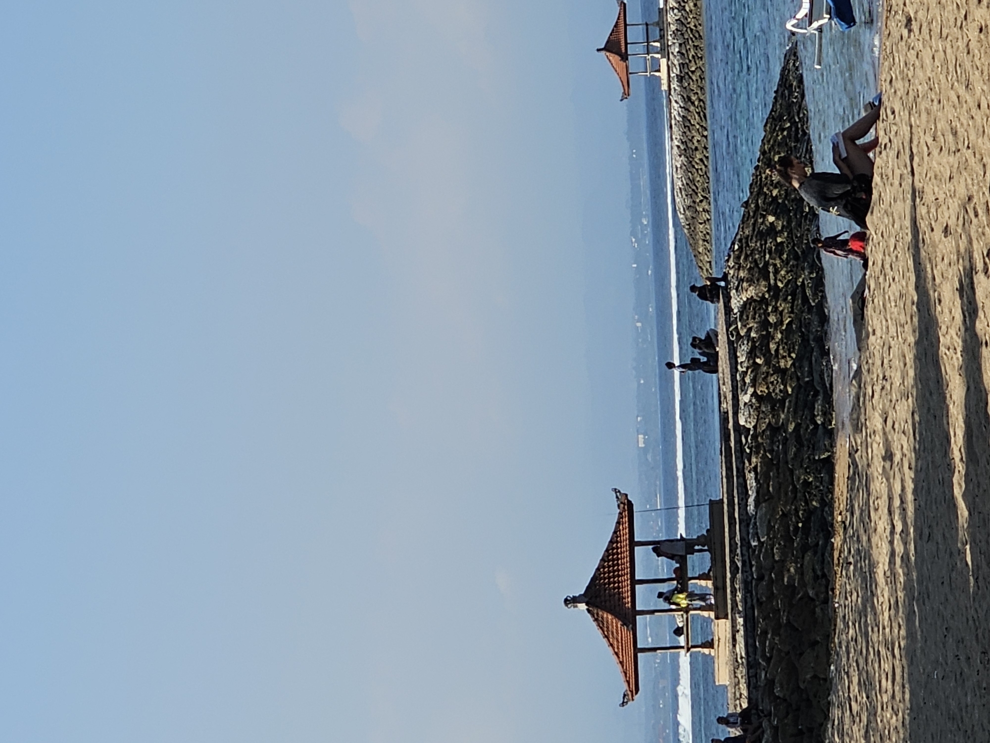 The image depicts a beach scene in Denpasar, Indonesia, likely Sanur Beach given the presence of traditional Balinese gazebos and stone jetties. In the foreground, a sandy beach stretches towards the water. A woman is seated on the sand, engrossed in reading a book, with a young child in a red skirt walking nearby. Further down the beach, a white boat is partially visible.
The middle ground features a rocky jetty extending into the blue ocean. On this jetty, a traditional Balinese gazebo with a red, multi-tiered roof provides shade for several people. Another, smaller gazebo is visible further out on a similar jetty. The water is a vibrant blue, with gentle waves lapping at the shore.
In the background, the coastline of Denpasar is visible, with distant hills and mountains forming a hazy silhouette against the clear blue sky. The overall impression is one of a tranquil beach day, with people enjoying the sun, sea, and shade of the gazebos.