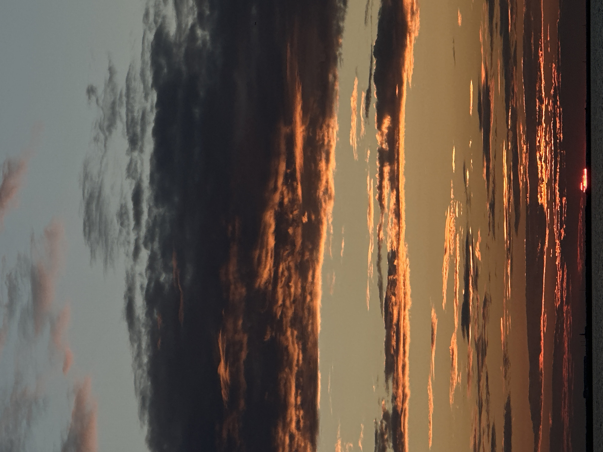The image depicts a sunset over a body of water, likely Lake Borovoye, near Kokshetau, Kazakhstan. The sky is filled with layers of clouds, ranging from dark, ominous formations to thin wisps illuminated by the setting sun. The lower clouds are a vibrant orange and red, indicating direct sunlight hitting them, while higher clouds are a mix of grey and pale pink. The horizon is barely visible as a dark line, suggesting it is the shoreline or a distant landmass. A small, red sliver of the sun is visible just above the horizon, casting a faint glow. There are no people or discernible objects in the immediate foreground, with the focus being entirely on the dramatic sky and the interplay of light and shadow. The overall atmosphere is one of natural beauty and tranquility.
