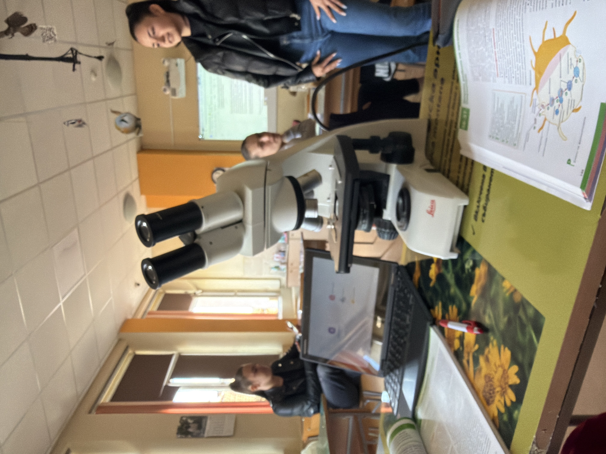The scene is a classroom in Panagyurishte, Bulgaria, likely a science lab. A white Leica microscope dominates the foreground, positioned on a desk with a laptop. A textbook showing an illustration of a cell structure is also on the desk. Two women are present; one is standing and dressed in a black jacket and jeans, while the other is sitting and also dressed in a black jacket. A third person, possibly a student, is visible in the background. The room features tiled walls, windows with closed blinds, and an overhead projector screen. The lighting appears to be a combination of natural and fluorescent.