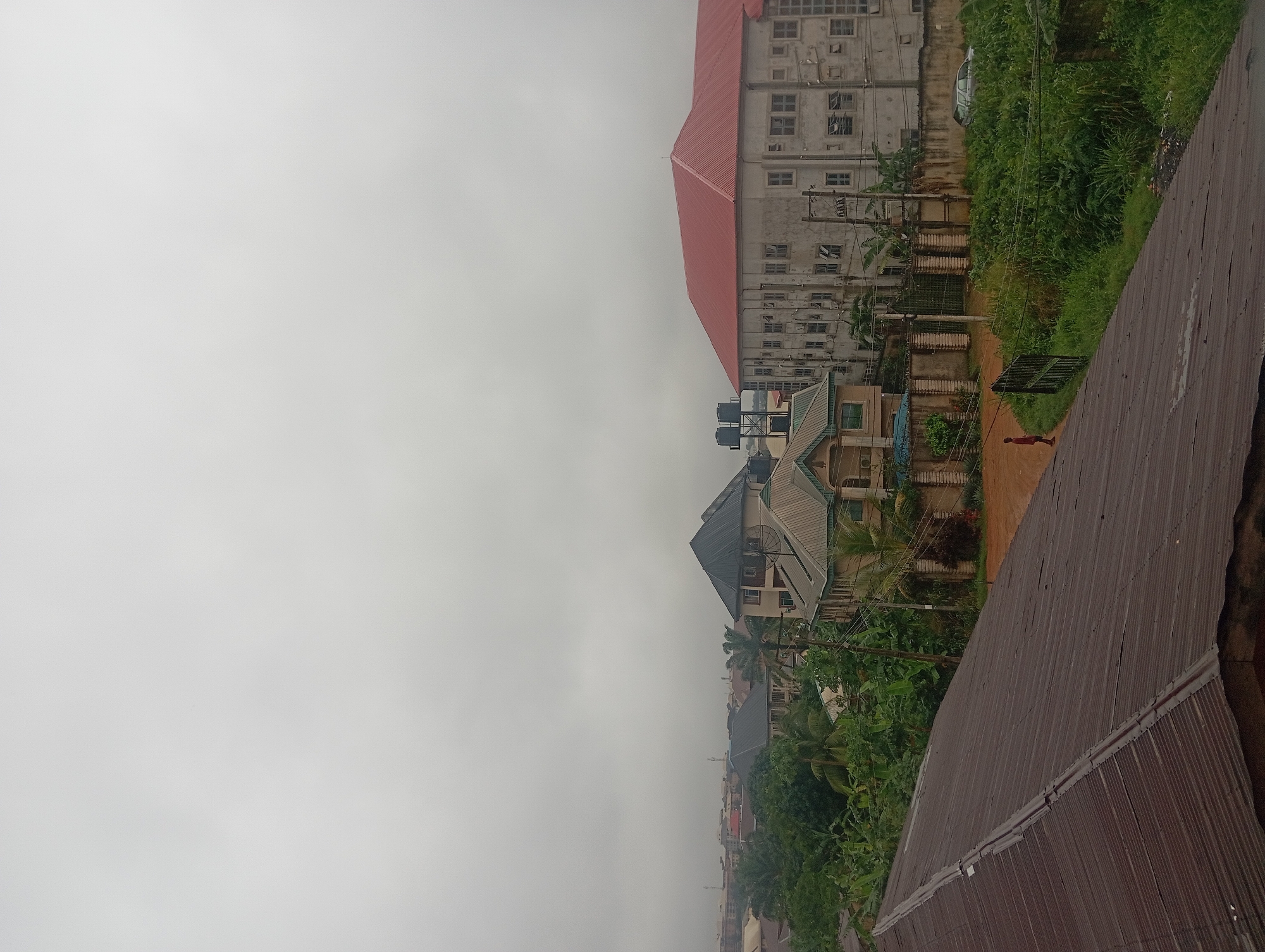 The image shows a cityscape under an overcast sky, likely taken from an elevated position. In the foreground, a weathered, corrugated metal roof dominates the lower left quadrant. Beyond this roof, several buildings are visible, varying in architectural style and height.
To the left and center, there are residential buildings with dark, sloped roofs and light-colored facades. Lush green vegetation, including trees and shrubs, surrounds these structures, suggesting a tropical or sub-tropical environment. Further in the background, a larger, lighter-colored building with a red, sloped roof is prominent. This building appears to have multiple stories and numerous windows.
A single individual, wearing a red shirt and dark pants, is standing on a dirt path near a fence in the middle ground. To the right, a patch of overgrown green grass and bushes is visible, with a light-colored car parked amongst the foliage. Electrical poles and wires are also scattered throughout the scene, indicating the presence of infrastructure. The overall impression is of a built-up urban or semi-urban area with signs of both residential and possibly commercial development, characterized by a modest density of buildings interspersed with natural greenery. The overcast sky suggests a cloudy or potentially rainy day.