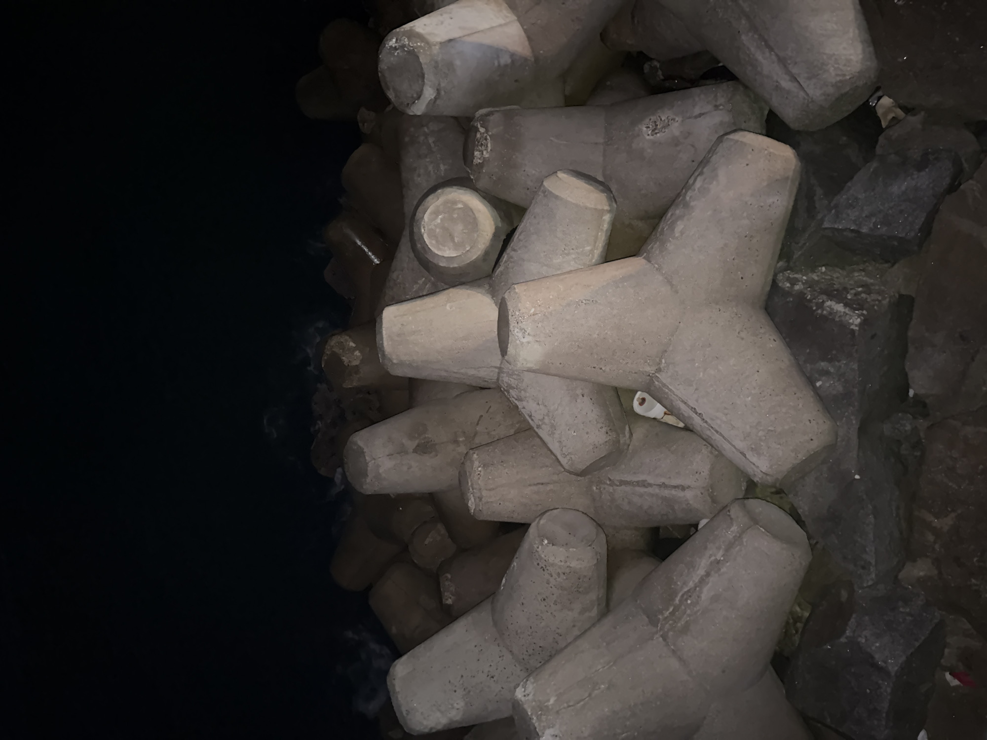 The image shows a cluster of concrete tetrapods on a rocky shoreline at night. The tetrapods are irregularly placed and appear to be part of a breakwater or seawall. The water is dark and appears calm, with small waves visible on the left side of the image. The sky is completely dark, indicating it is nighttime. The lighting suggests artificial illumination from an unseen source, casting shadows and highlighting the textures of the concrete and rocks. There are no people or animals visible in the image. The scene is likely located at a coastal area in Gangneung-si, South Korea, given the context.