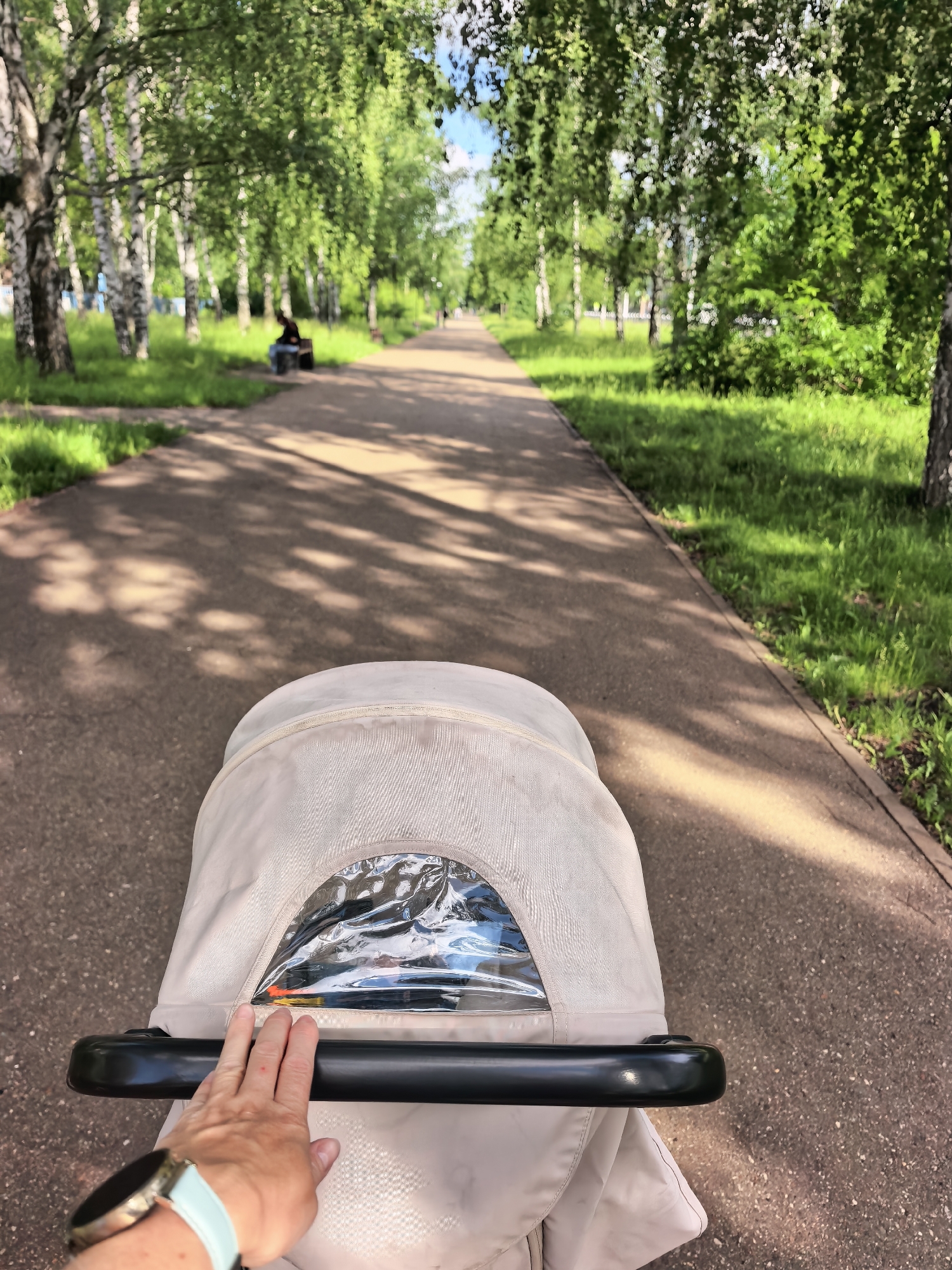 The image depicts a first-person perspective looking forward over a beige baby stroller on a paved pathway within a park in Sterlitamak, Russia. A left hand, wearing a smartwatch with a light blue band, rests on the black handlebar of the stroller, which features a clear plastic viewing window in its canopy. The asphalt pathway extends into the distance, bordered by vibrant green grass and numerous birch trees with white bark and lush green leaves. Sunlight filters through the tree canopy, casting prominent dappled shadows on the pathway. In the midground, to the left of the path, a single individual is seated on a brown park bench under the trees, clad in dark attire. The background consists of more trees and greenery, indicating a continuous park environment under a bright sky.