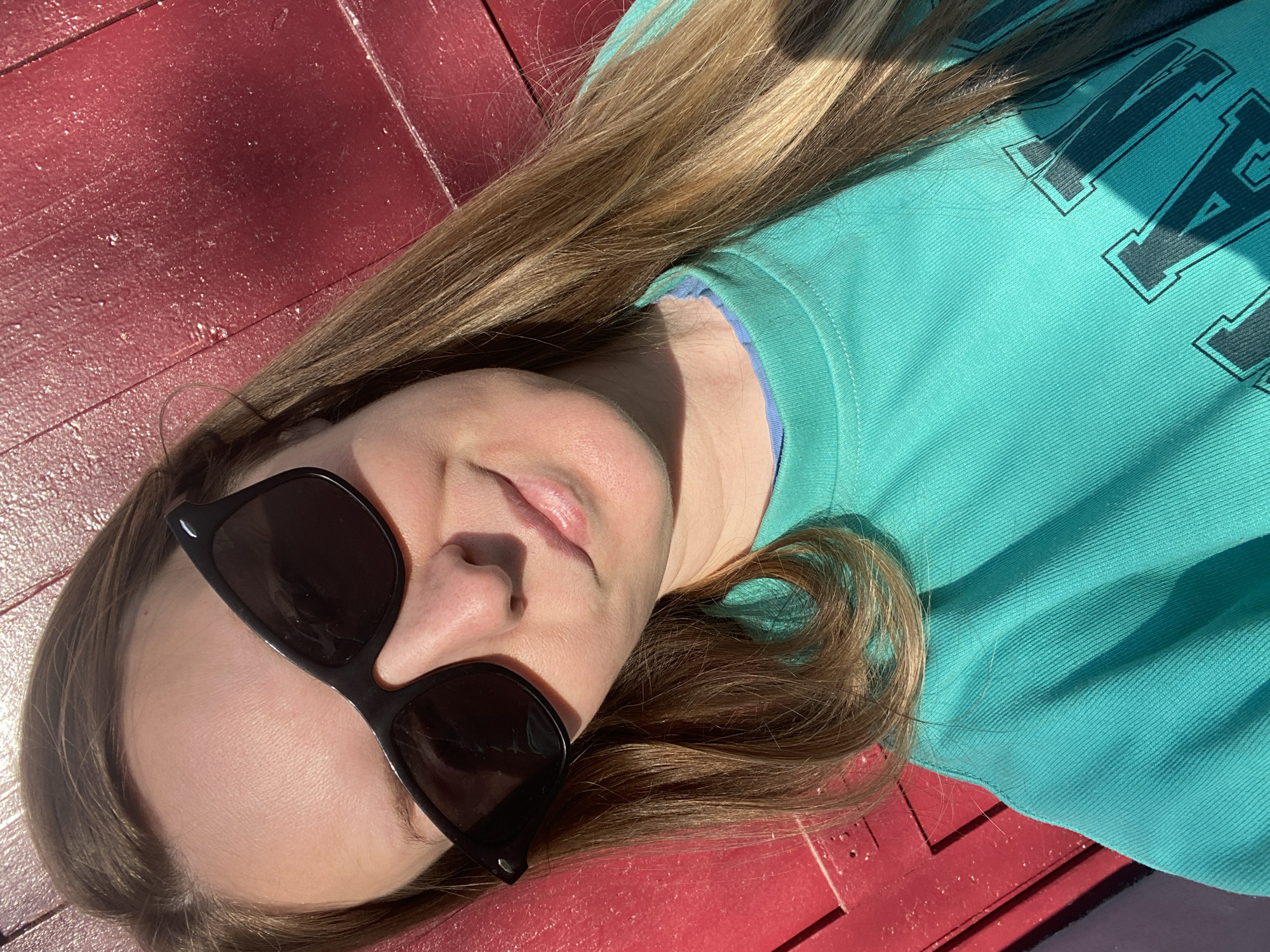 A woman lies on a red wooden surface. She is wearing black sunglasses and a teal sweatshirt with dark lettering. Her hair is long and brown. The location is unknown and cannot be determined from the image.