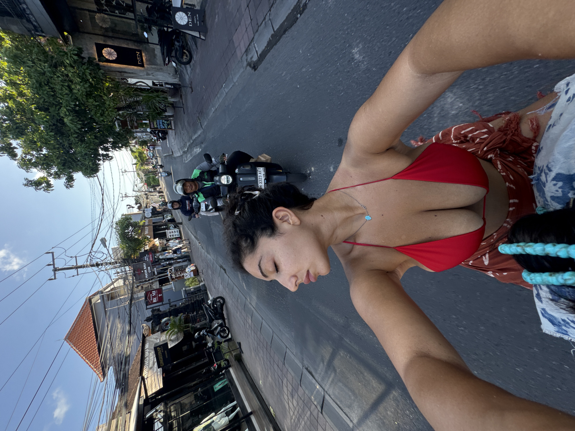 The image shows a woman with dark hair and a red bikini top riding on a motorbike in North Kuta, Indonesia. The woman is looking down, towards the front of the bike. The streetscape includes shops and other motorbikes, with signs visible for 'PUCUK BALI GYM COFFEE,' 'BUMI,' 'GINGERSNAP,' 'RUNG BU MI' and 'DEN.' The license plate of a motorbike is partially visible and reads 'K 3323 AET.' There are power lines and trees along the street, with a blue sky overhead.