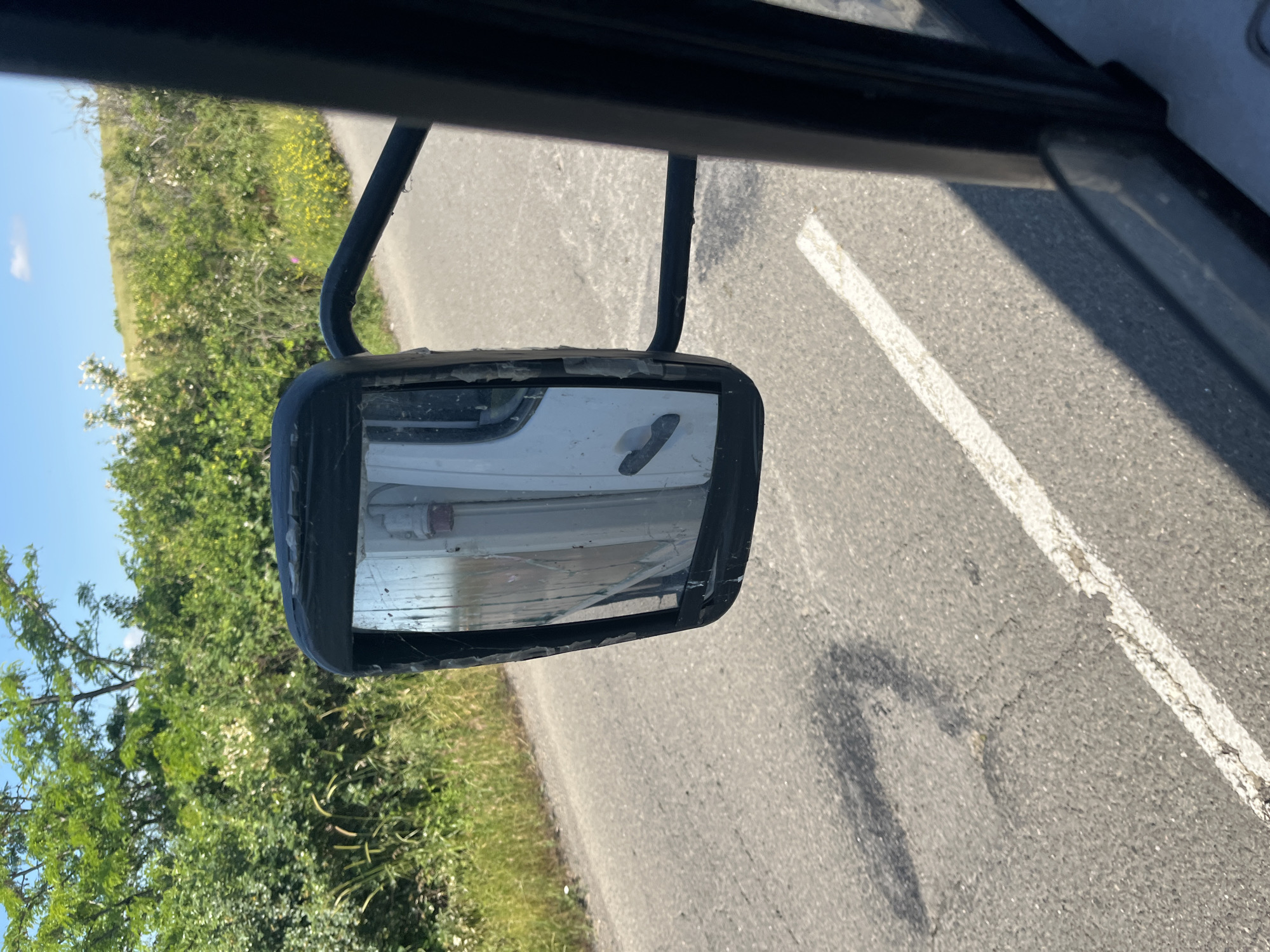 The image is a close-up, right-side exterior shot of a white vehicle, likely a van or truck, seen through its side mirror and window frame. The scene appears to be on a rural road in Tankovo, Bulgaria. The side mirror, with its black casing, is prominently featured, reflecting the side of the vehicle, a white door handle, and a vertical red and grey electrical outlet or connector. The mirror housing shows signs of wear and possibly some damage or disrepair, with visible tape or adhesive.
Beyond the vehicle, a paved road with a broken white line runs alongside a vegetated embankment. Lush green bushes and trees line the embankment, with some scattered yellow wildflowers and a single pink flower visible. The sky is blue with a few wispy white clouds. The overall impression is one of a sunny day in a rural setting, possibly during the summer months given the vibrant green foliage. The perspective suggests the photograph was taken from within the vehicle, looking outwards and into the side mirror. There are no people visible in the image.