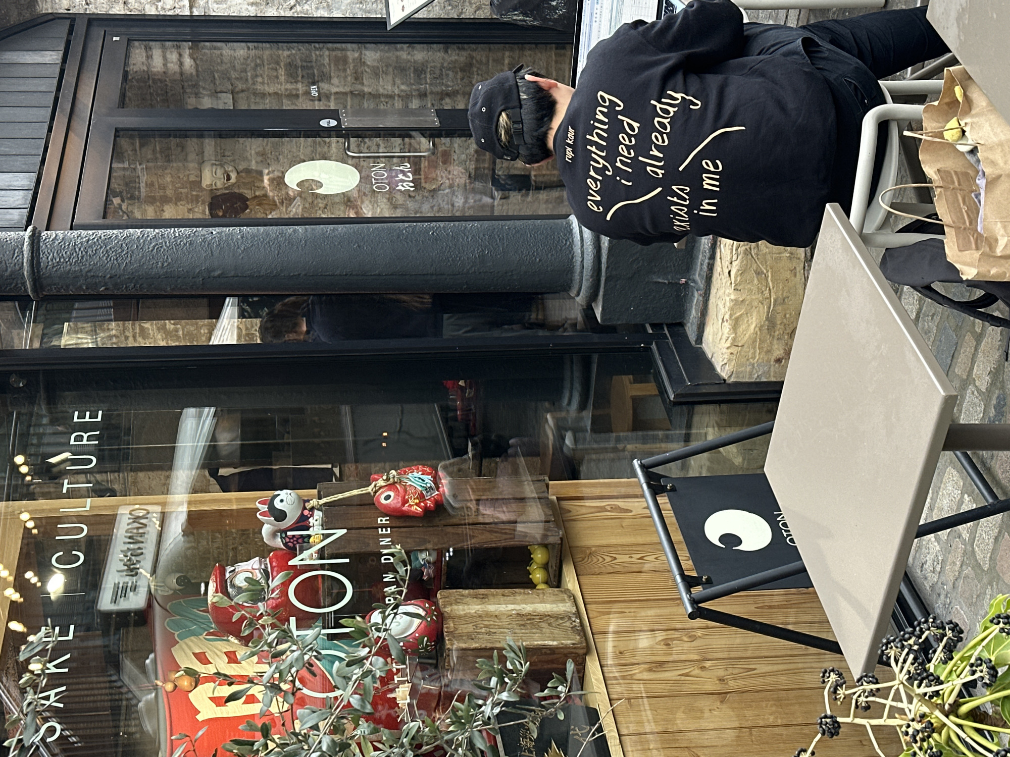 The image captures an outdoor scene, likely in London, United Kingdom, featuring a restaurant named "OTON". The restaurant's window display includes sake and cultural items. Outside, a person wearing a black hoodie and a black baseball cap is seated at a table, working on a laptop. The hoodie has the text "rupi kaur everything i need already exists in me" printed on the back. The table is light brown with a black frame. A paper bag sits on the chair next to the person. The building exterior appears to be brick. The setting is a blend of traditional architecture and modern dining.