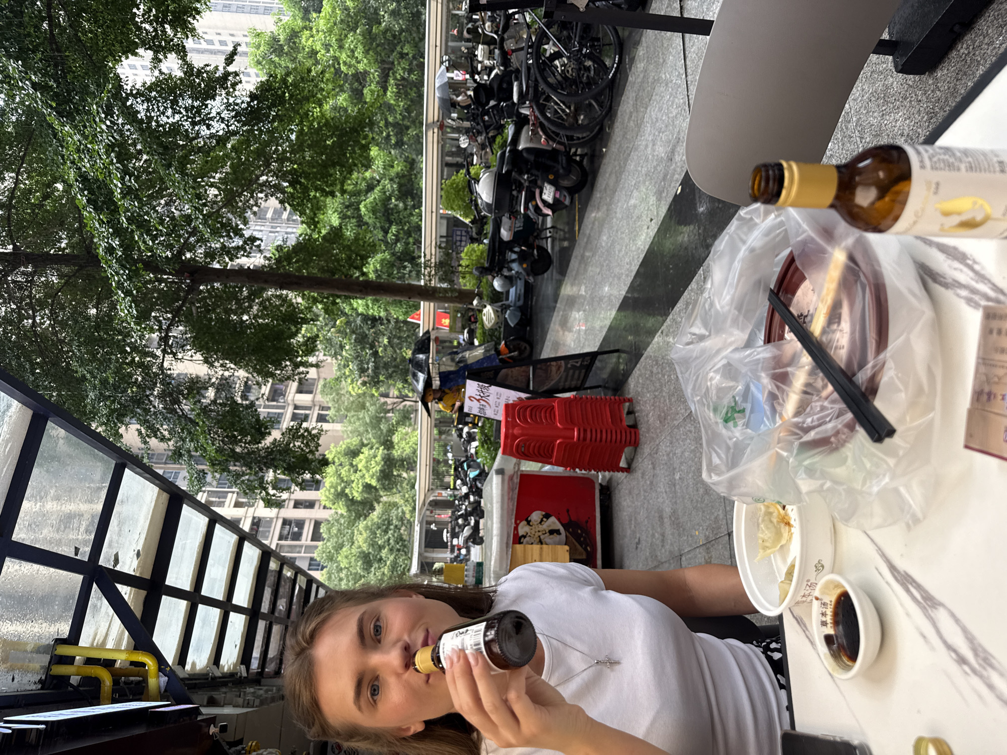 In Haizhu District, China, a young woman with blonde hair is seated at an outdoor table, holding a small bottle to her lips as if drinking. She is wearing a white t-shirt and a silver cross necklace. In front of her, on the table, are a small dish of soy sauce, a bowl of dumplings, chopsticks, and a plastic bag containing more food. A partially visible open bottle with a golden cap stands next to the food. The background shows a street scene with parked motorcycles, a red stack of chairs, a food stall, and tall buildings amidst lush green trees. The sky appears overcast, and the ground is wet, suggesting recent rain.