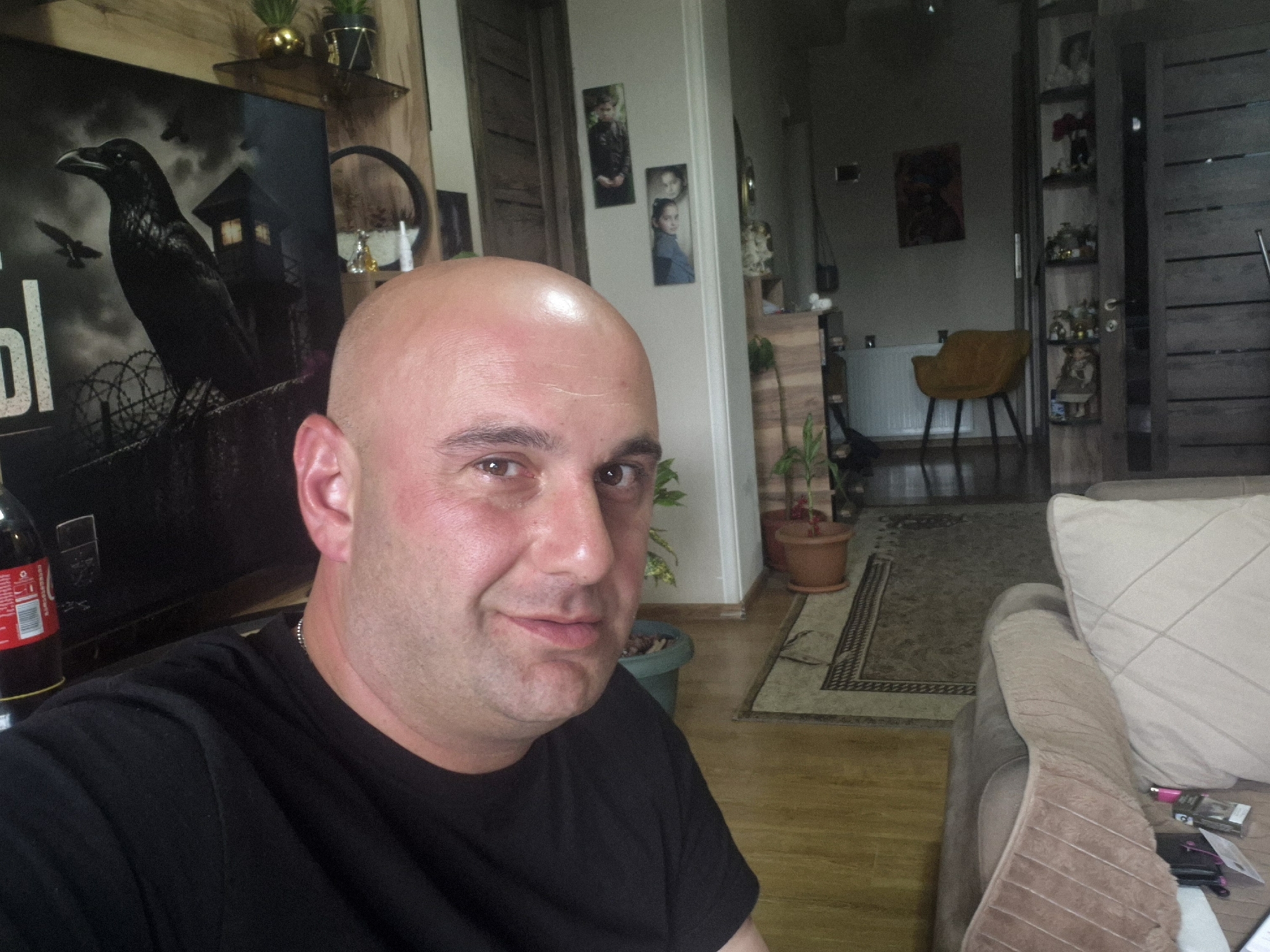 A bald man wearing a black t-shirt is in the foreground of a room, possibly in an apartment or house. Behind him is a poster featuring a black crow and a building with barbed wire. In the background, there is a doorway leading to another room with a yellow chair and a bathtub visible. A decorative shelf unit with various ornaments can be seen to the right of the doorway. Plants in terracotta pots are positioned near the doorway. Further details include personal pictures on the wall and a sofa in the foreground. The location appears to be Tbilisi, Georgia, based on the architectural features and decor potentially associated with residential spaces in the city.