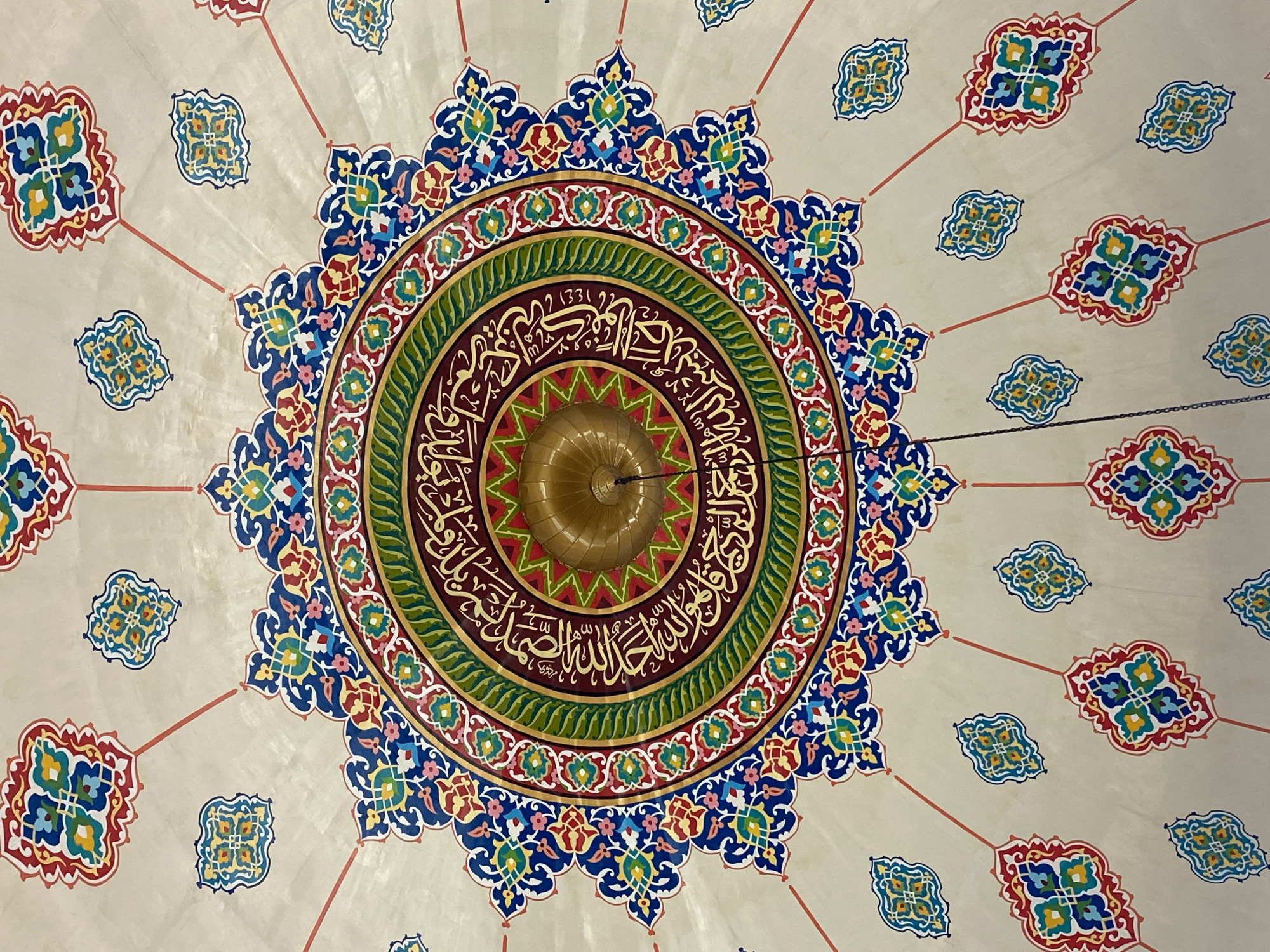 The image shows a close-up view of a highly ornate ceiling in a building, likely a mosque, due to the Islamic calligraphy and geometric patterns. The ceiling is painted with vibrant colors including red, blue, green, yellow, and gold. In the center, there is a golden dome-like fixture with intricate carvings, and surrounding it is a circular band of Arabic script. Radiating outwards from the center are floral and geometric motifs, with smaller decorative elements scattered across the pale ceiling. The overall impression is one of grandeur and detailed craftsmanship. This ceiling is located in the city of Lhokseumawe, Indonesia.