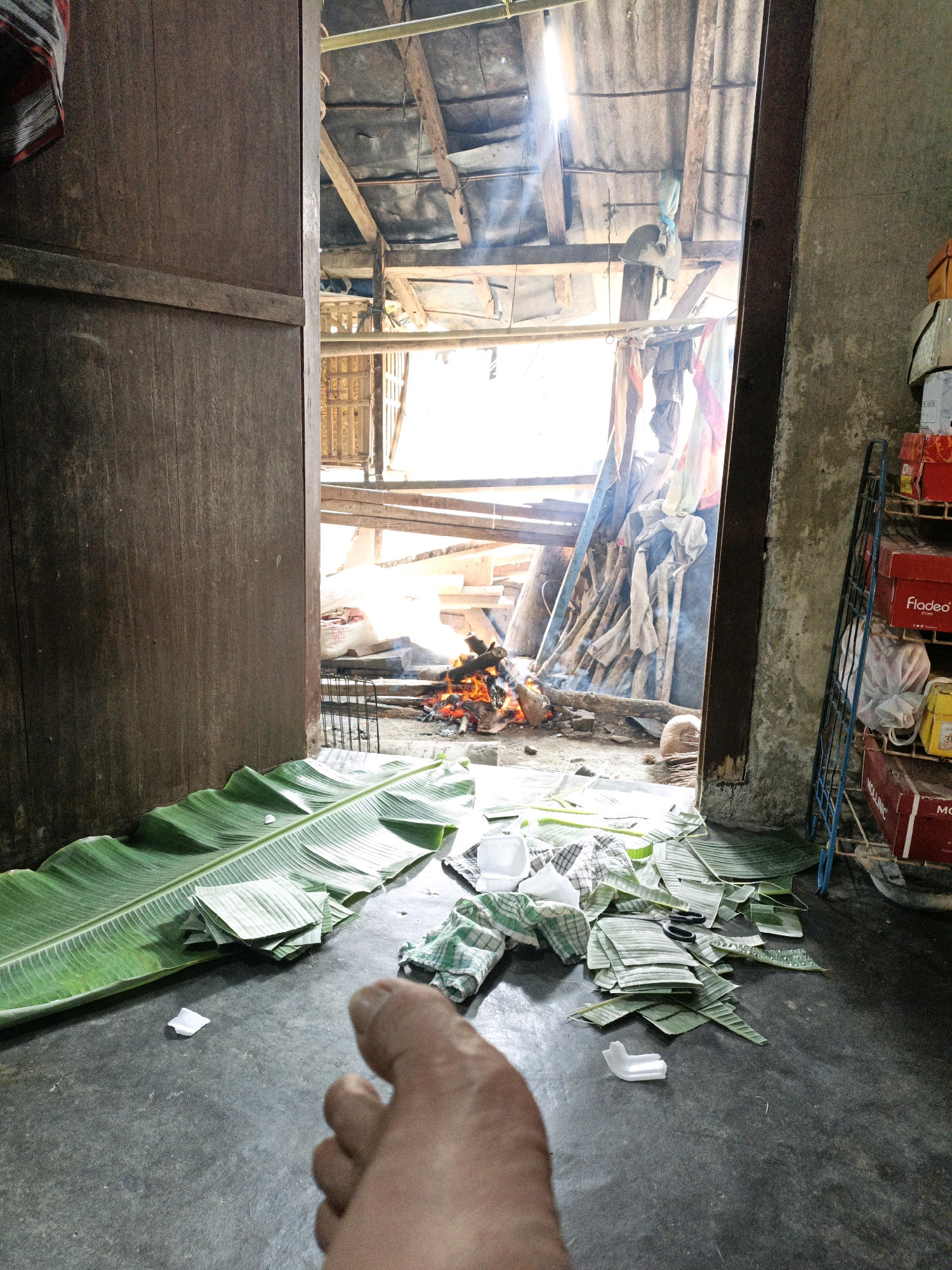 The image depicts an indoor scene with an open doorway revealing an outdoor area where a fire is burning. The interior features a rough, concrete floor. To the left, a large, wooden door stands open. In the foreground, several large, green banana leaves are spread on the floor, along with a pile of smaller, cut pieces of banana leaves and a checkered cloth. A human foot is visible in the lower right corner.
The doorway opens to a rustic outdoor space with a corrugated metal roof supported by wooden beams. Debris, including wood and fabric, is scattered around. The fire pit is visible, with flames and smoke rising. A metal grill is positioned near the fire. In the background, stacks of boxes are visible on a blue metal shelf unit against a rough concrete wall.
Based on the context provided, this scene is located in Bunut, Indonesia. The overall impression is one of a utilitarian or possibly makeshift cooking or preparation area, with evidence of recent activity involving food preparation using banana leaves and an open fire.