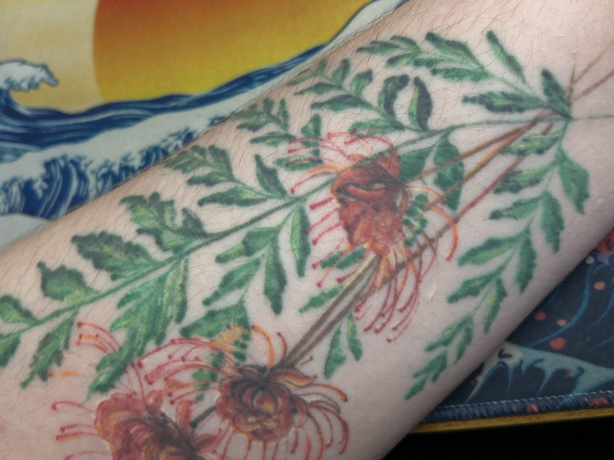 The image shows a close-up of a forearm with a tattoo depicting red spider lilies (Lycoris radiata) and foliage in green ink. The skin is light-toned with visible hair follicles. The background includes a partially visible mousepad with a print resembling Hokusai's "The Great Wave off Kanagawa" and a golden sun. The location in the city of Ural'skoe, Russia, is not determinable from the image alone.
