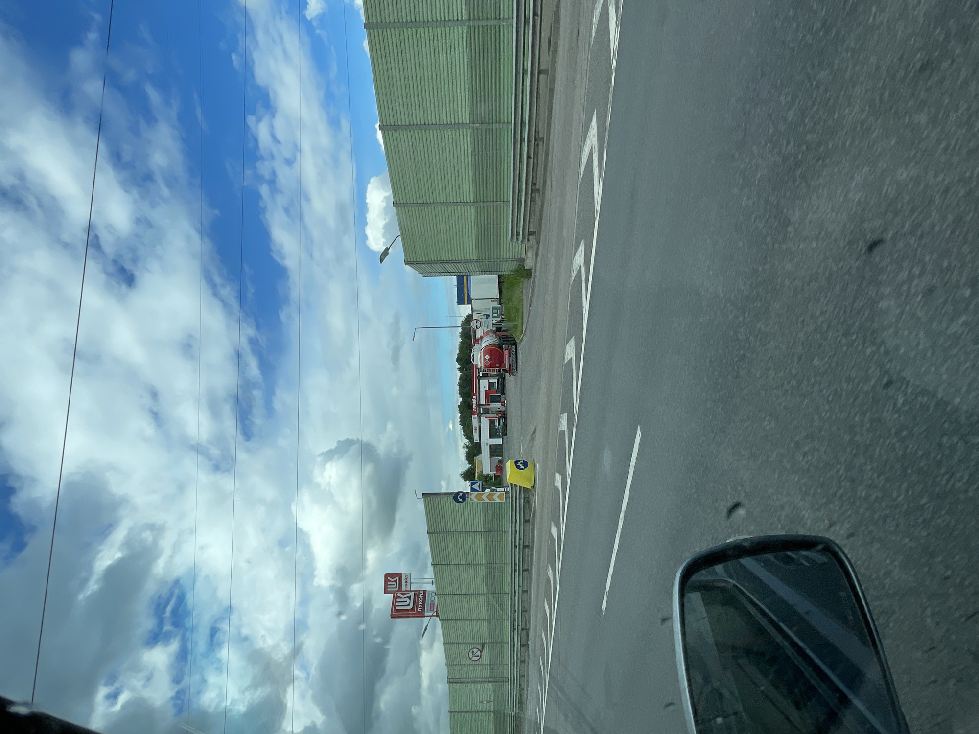The image shows a view from inside a vehicle on a road in Berezhki, Russia. A Lukoil gas station with multiple fuel trucks is visible along the side of the road, separated from the lanes by a tall green noise barrier. Road signs are also present. The sky is partially cloudy with power lines visible. A side mirror of the vehicle is visible in the bottom right.