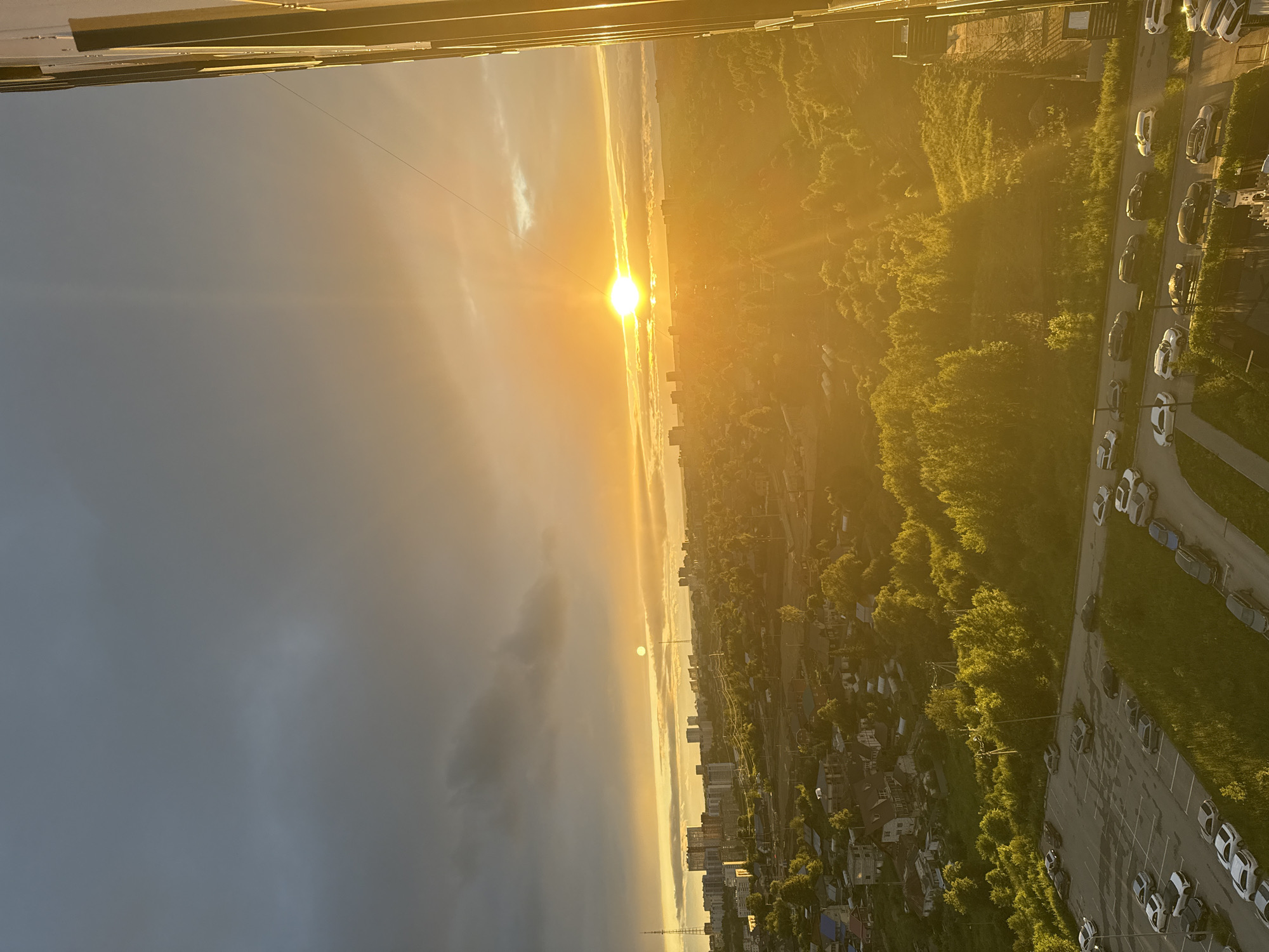 The image depicts a panoramic view of a city during sunset, likely in Perm, Russia. The sun is low on the horizon, casting a warm, golden glow across the scene and creating a dramatic sky with streaks of orange, yellow, and grey clouds. The light dramatically illuminates the landscape, highlighting the vibrant green of trees and the tops of buildings.
In the foreground, the edge of a building or balcony is visible, suggesting the photo was taken from a high vantage point, possibly an apartment building. Below, a dense urban area unfolds, characterized by a mix of apartment buildings and smaller houses, interspersed with lush green trees. A road lined with parked cars runs through the scene, suggesting a typical urban environment. The overall atmosphere is one of tranquil beauty, with the setting sun bathing the city in a serene, ethereal light. The time of day is clearly sunset, with the sun's low angle and the intense golden light being key indicators. The weather appears to be clear with some cloud cover, contributing to the dramatic sky. The presence of urban architecture and the general landscape strongly suggest a city environment, consistent with the provided location context of Perm, Russia.