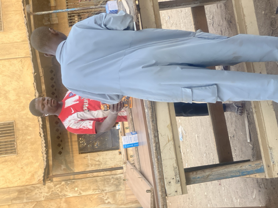 Two men stand outdoors near a workbench. One man wears a light blue jumpsuit and faces away from the camera. The other wears a red and white athletic shirt and holds a drill. The workbench is surrounded by wooden planks. Boxes are placed on the bench. A building with a window and metal grate is in the background. The ground is unpaved.