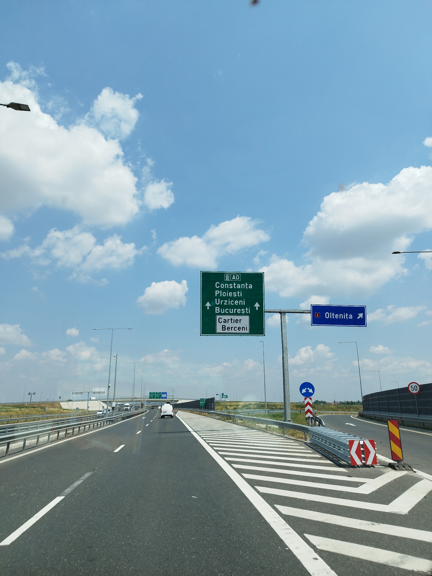 The image shows a highway in Popești-Leordeni, Romania. A green highway sign indicates directions to Constanța, Ploiești, Urziceni, București, and Cartier Bêrceni. A blue sign points towards Oltenița. The highway is paved and has white lane markings. There are guardrails along the side of the road, and several vehicles are visible on the highway, including a white van. The sky is blue with white clouds. There are streetlights and traffic signs visible along the road. A speed limit sign of 50 is also visible. The scene appears to be a sunny day.