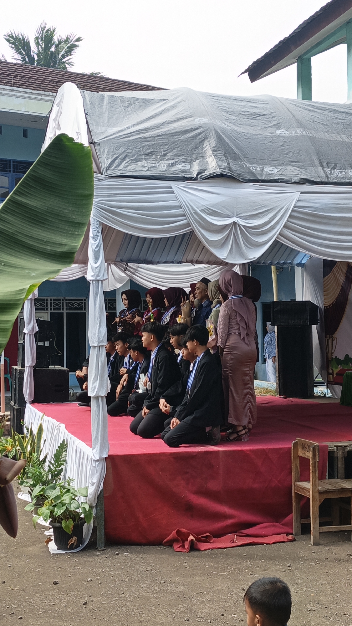 In Tigaraksa, Indonesia, a group of students, some in graduation attire with medals, are seated on a raised red-carpeted stage. The stage is covered by a white tent with draped fabric. Several adults, some wearing hijabs, are standing behind the seated students. One adult woman, in a long, form-fitting dress and hijab, stands towards the right side of the stage, facing away from the camera. A wooden chair is placed to the right of the stage. In the foreground, a child with dark hair is visible from the shoulders up, facing away from the camera. The background shows a building with blue walls and a brown roof, and a palm tree. The ground surrounding the stage is made of gravel and dirt.