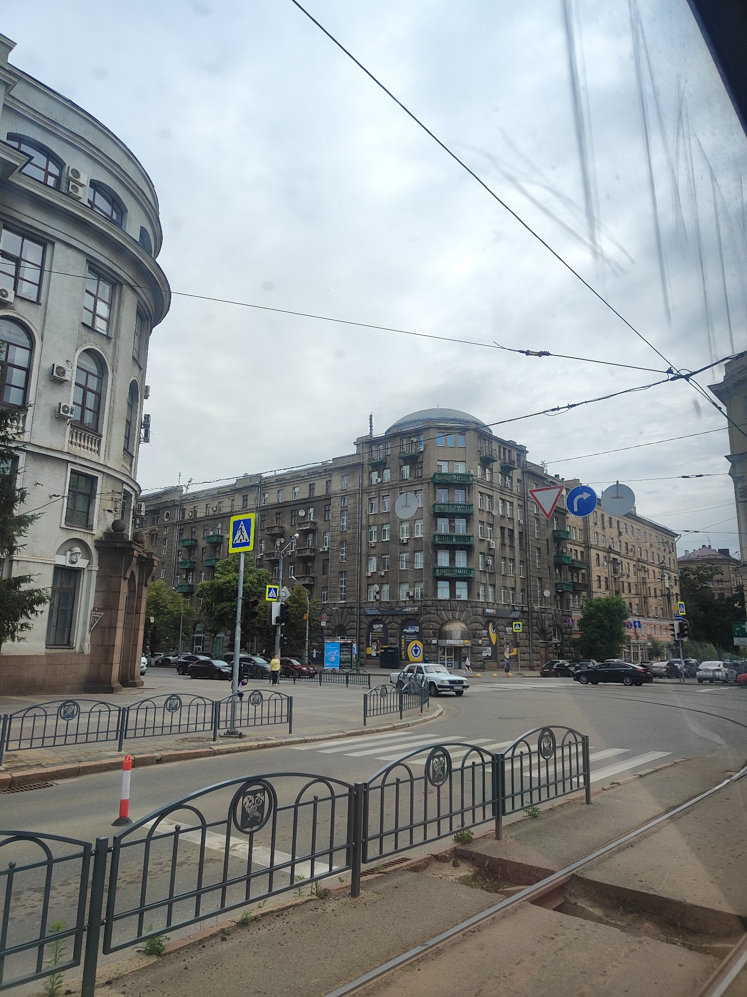 The image shows a street scene in an urban environment, likely during daytime with overcast skies. On the left, a large, curved, grey building with arched windows and balconies is prominent. In the center and right, a series of older, multi-story apartment buildings with a similar architectural style are visible, featuring numerous windows and balconies. The street is paved, with trolley tracks running through it. There are several cars on the road, some parked and others in motion. Pedestrian crossings are marked on the road, and traffic signs, including a pedestrian crossing sign and directional signs, are present. A few people are visible on the sidewalks and crossing the street. Metal fences with decorative elements line the sidewalks. Overhead, power lines crisscross the sky, suggesting the presence of public transportation like trams or trolleybuses. The overall impression is that of a busy city intersection.