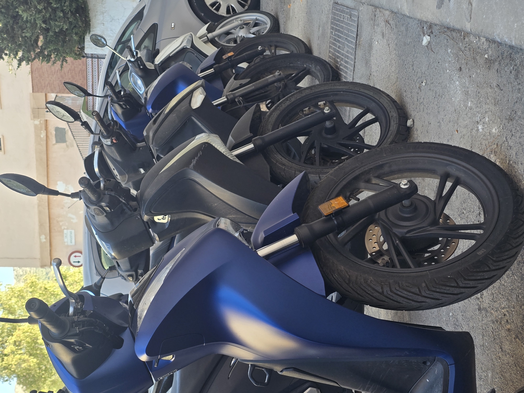 A row of parked scooters is seen from a low angle, with the closest scooter being a vibrant blue. Behind it, other scooters in black and white are also parked. A grey car is visible in the background. The scene is set on a street in the city of Málaga, Spain, as indicated by a street sign that reads "CALLE ANGUISTAS". The ground is paved with concrete, and a metal grate is present on the right side of the image. The lighting suggests it is daytime, with sunlight casting shadows and highlighting the metallic surfaces of the scooters. There are no people visible in the frame. The scooters appear to be stationary, parked along the side of the street.