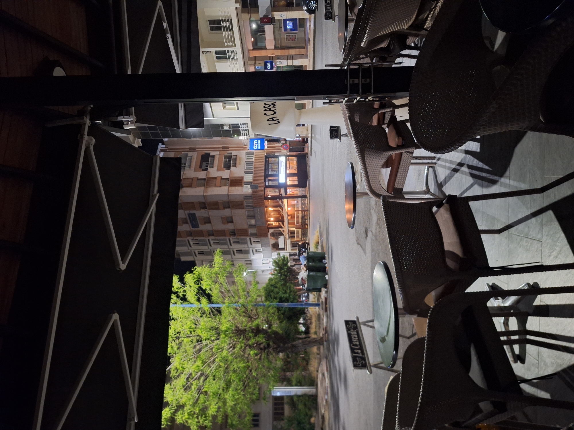 The image depicts an outdoor seating area of a restaurant in Safi, Morocco, at night. The restaurant, "La Cascade," is evident by the signage on a table. Wicker chairs surround small tables on a paved surface. In the background is a street with lit buildings, including "LA CASCO," and trees lining the roadside. Trash cans are visible further down the street. The perspective is from under an awning-like structure of the restaurant.
