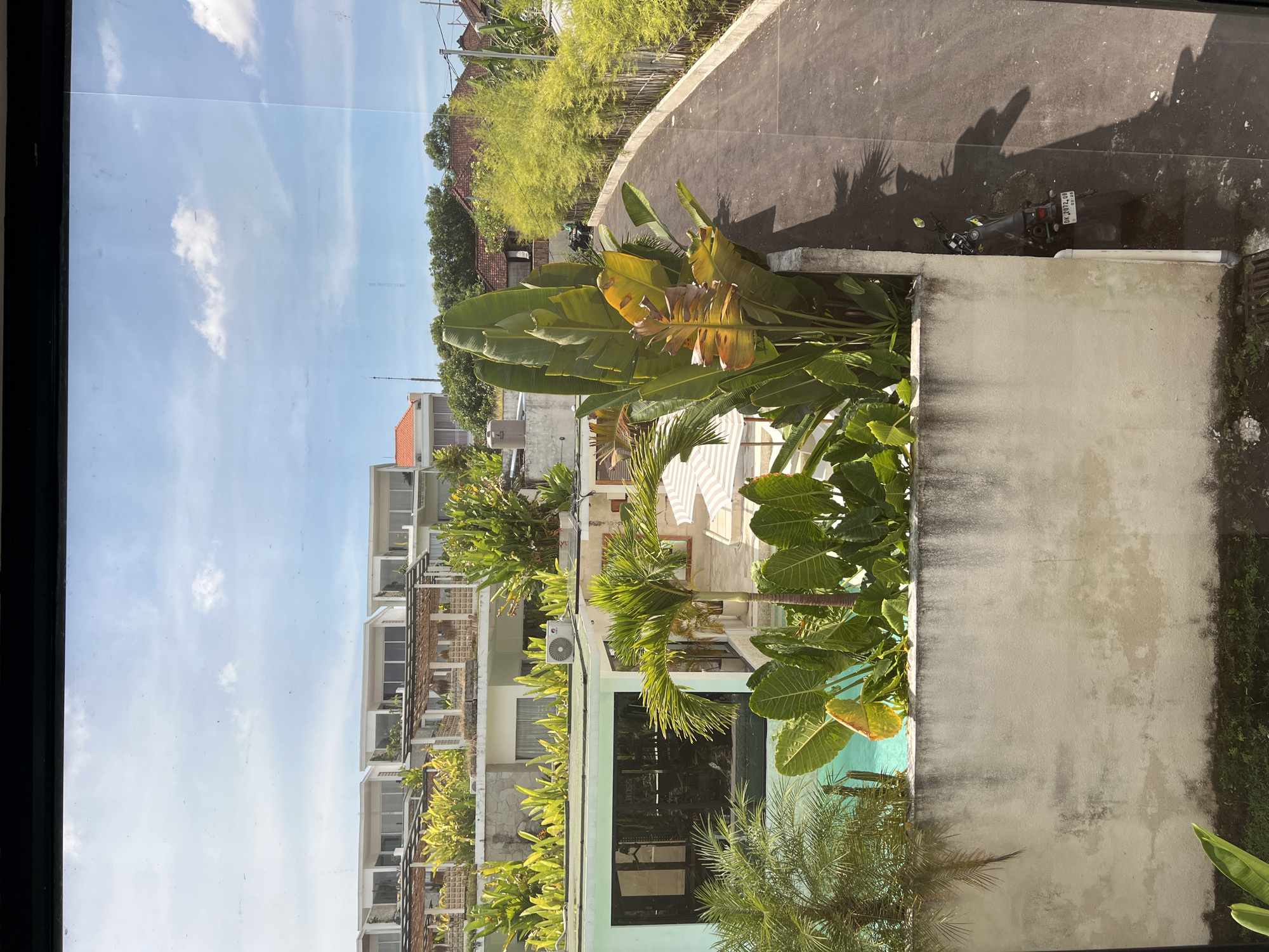The image shows an outdoor scene in Buduk, Indonesia, as viewed from a high vantage point, possibly a window. The sky is visible with light clouds. Buildings with multiple balconies and greenery on each level are seen to the left. Lush tropical plants are prominent in the midground, including banana trees and palm trees, partially obscuring what seems to be a swimming pool. A gray concrete wall is on the right. Further up, a paved road, likely for vehicles, runs parallel to the scene, and a parked motorbike is visible on the road. Above the road, there are additional buildings obscured by more dense tropical vegetation.