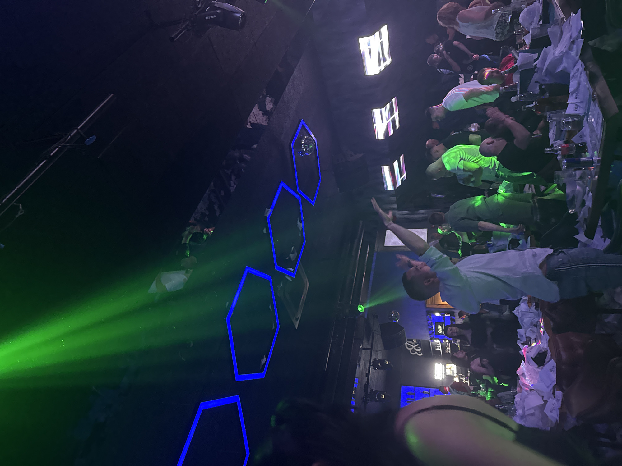 The image depicts a crowded indoor entertainment venue, specifically "MAMA's Place," located in Sofia, Bulgaria, as indicated by visible branding on table items. The scene is illuminated by artificial lighting, predominantly green spotlights creating visible beams from the ceiling, and blue hexagonal LED outlines on the dark ceiling. A small disco ball is suspended within one of these hexagonal fixtures. White rectangular light panels are visible on the far right ceiling.
Approximately 30-40 individuals are present, consisting of men and women of various ages, primarily adults. Many are standing, some with raised arms, suggesting dancing or cheering. In the mid-ground, a man in a light-colored shirt is captured with both arms extended upwards. Other individuals are seated at dark wooden tables in the foreground and mid-ground. These tables are extensively covered with numerous crumpled white papers or napkins. Table contents include various bottles (alcoholic spirits, soda), drinking glasses, and ice buckets. A person with dark hair, seen from the back, is visible in the foreground left. In the background left, a bar area is distinguishable by blue backlighting and shelves holding bottles, alongside a partial sign displaying an ampersand and possibly a letter 'B' or 'R'. The floor is largely obscured by tables and people. The overall environment is dark, suggesting a nightclub or event space atmosphere.